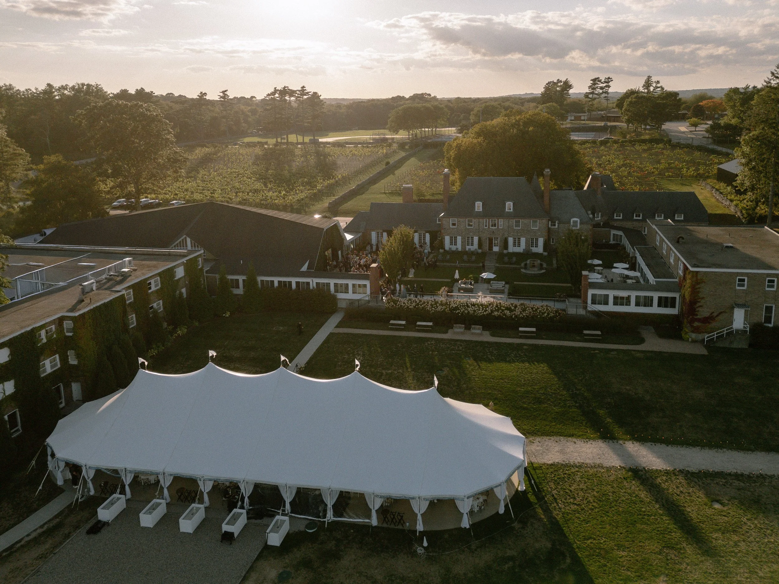 Aerial view of an elegant wedding venue featuring a large white sailcloth reception tent, historic estate buildings, landscaped grounds, and surrounding vineyards at sunset.