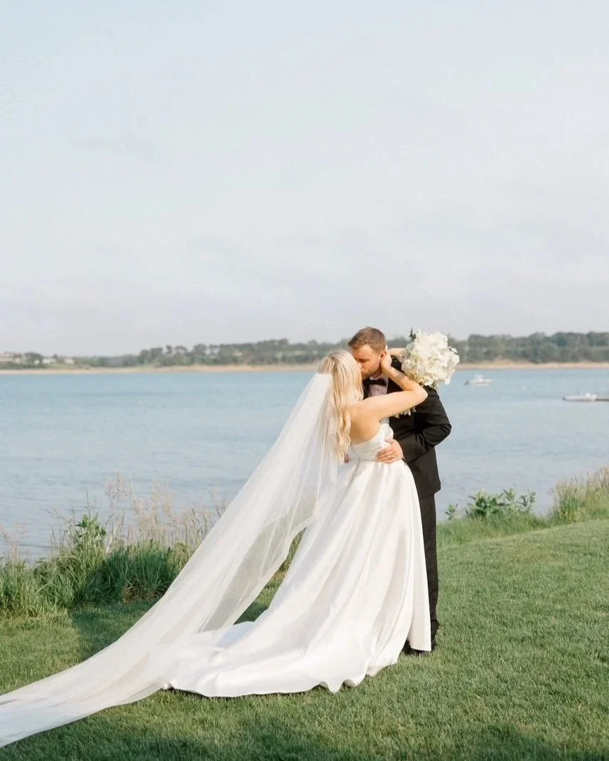 Bride and groom kiss by the water, with a flowing veil and waterfront backdrop.