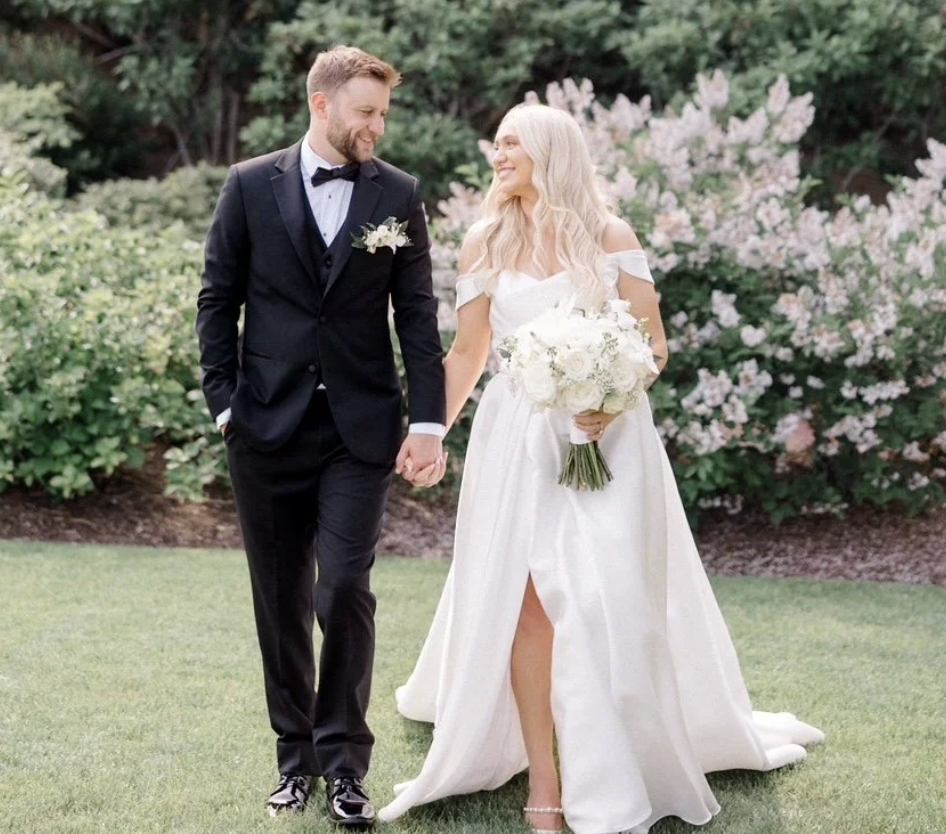 Bride and groom walk hand in hand through a garden after their wedding ceremony, smiling at each other while the bride carries a white bouquet.