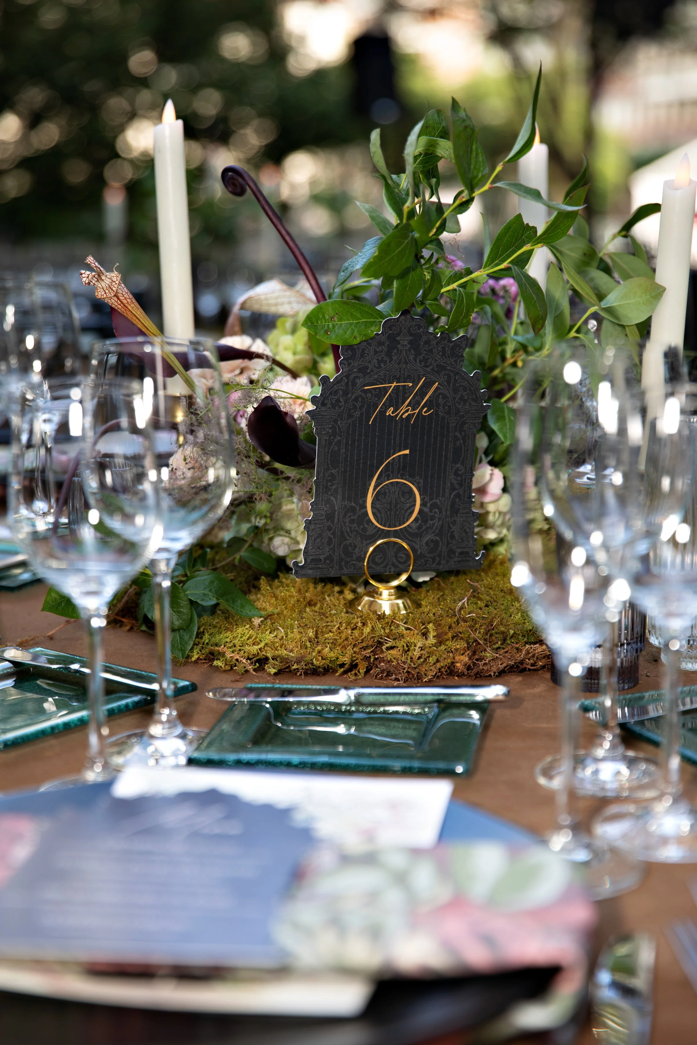 A formal outdoor dining table with a centerpiece of greenery, flowers, and candles, set for an event. The table has a black place card with gold writing indicating 'Table 6' and is surrounded by multiple wine glasses and plates.