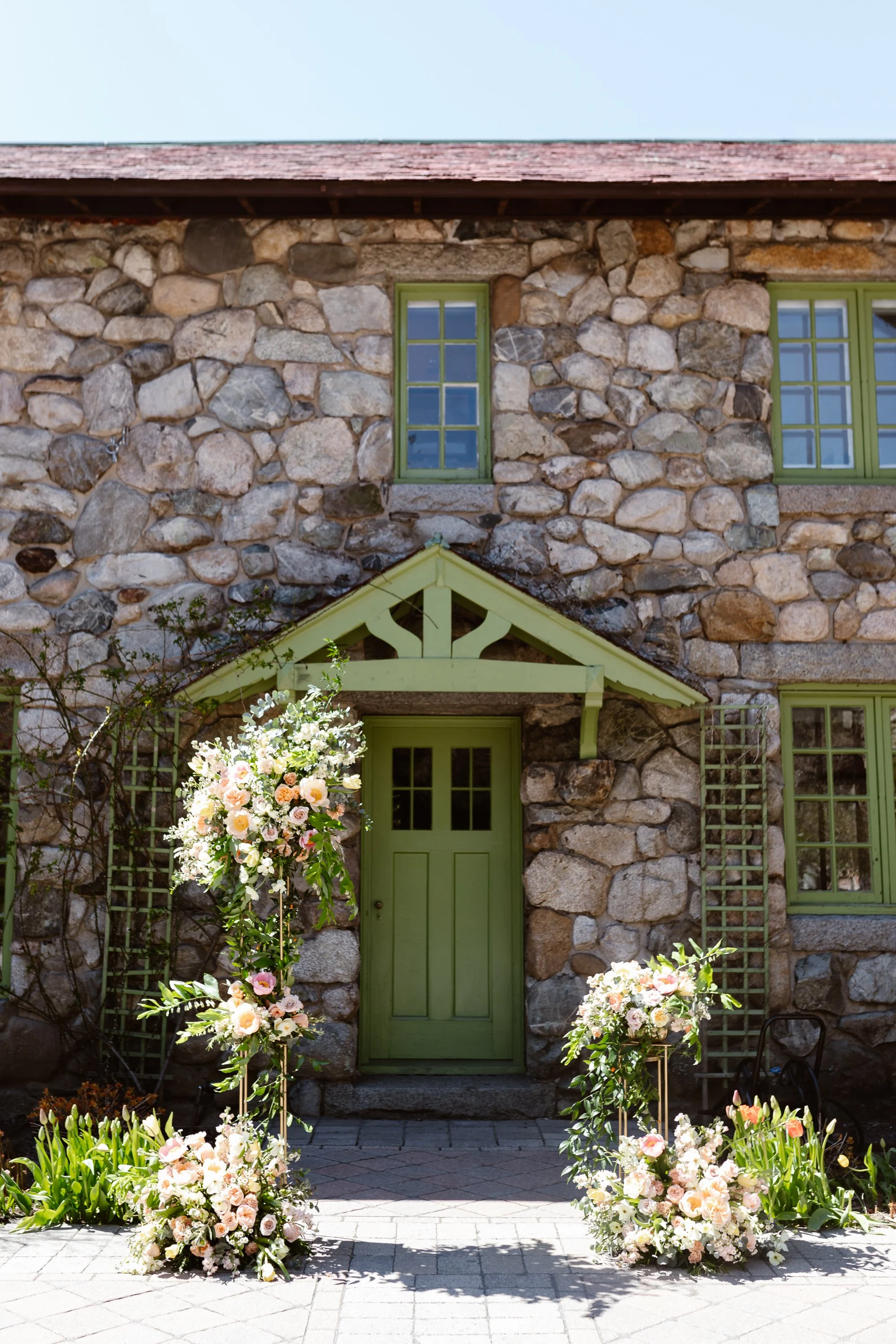 Decorated stone house entrance with green door and windows, featuring floral arrangements with pink and white flowers and greenery.