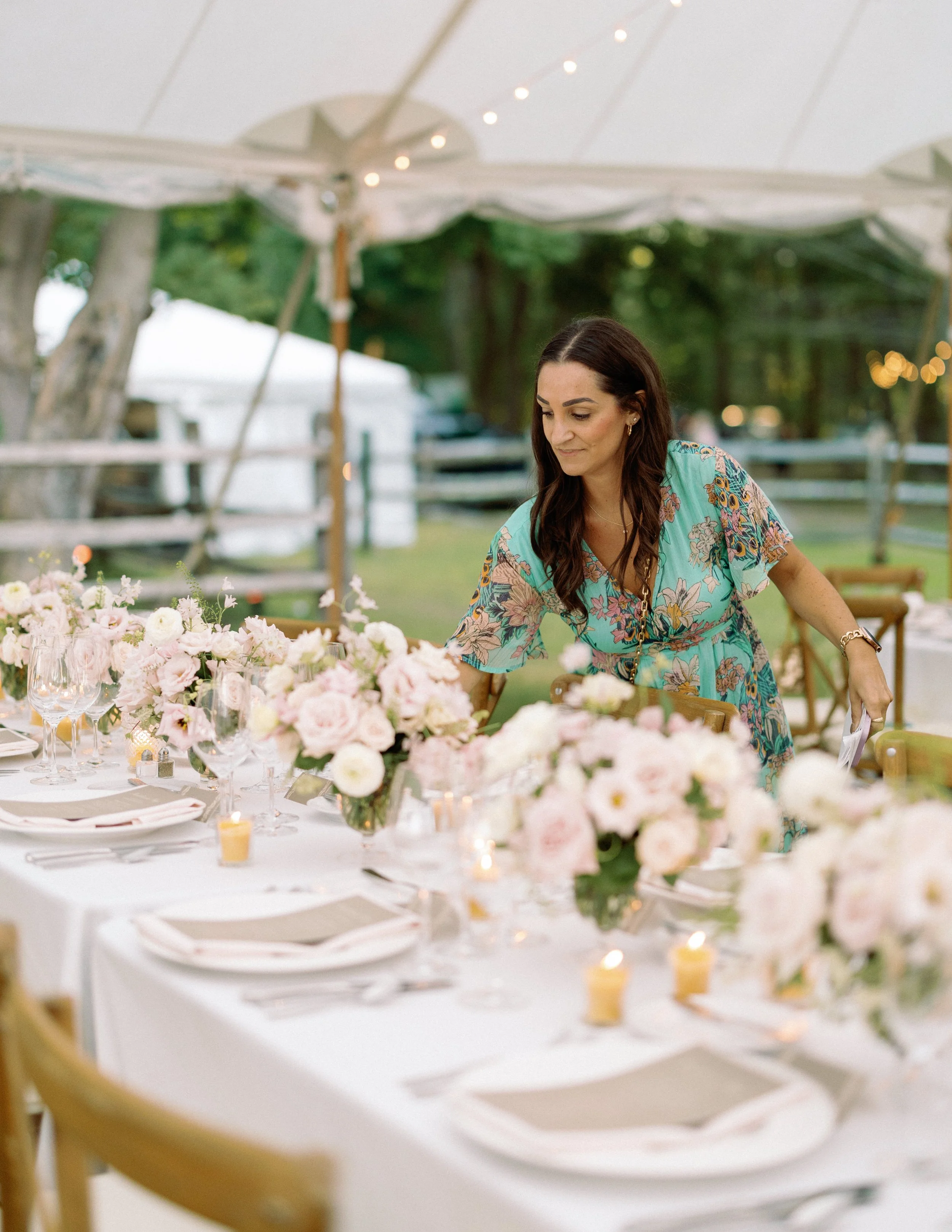 Wedding planner styling an elegant reception table beneath a sailcloth tent with blush floral centerpieces, candlelight, and refined place settings before guests arrive.