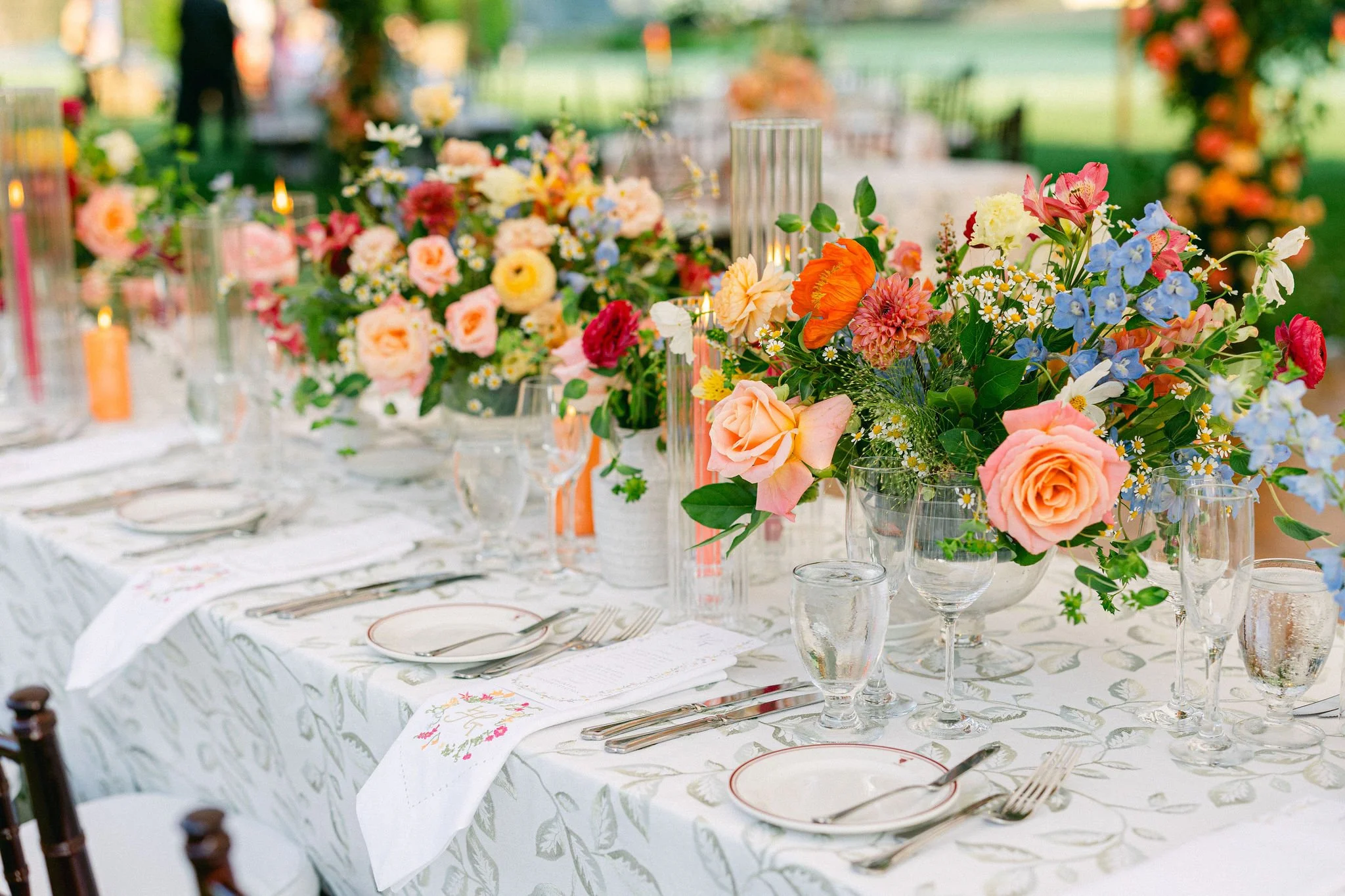 A beautifully decorated outdoor banquet table with elegant floral centerpieces, glassware, plates, silverware, and white napkins with floral embroidery.