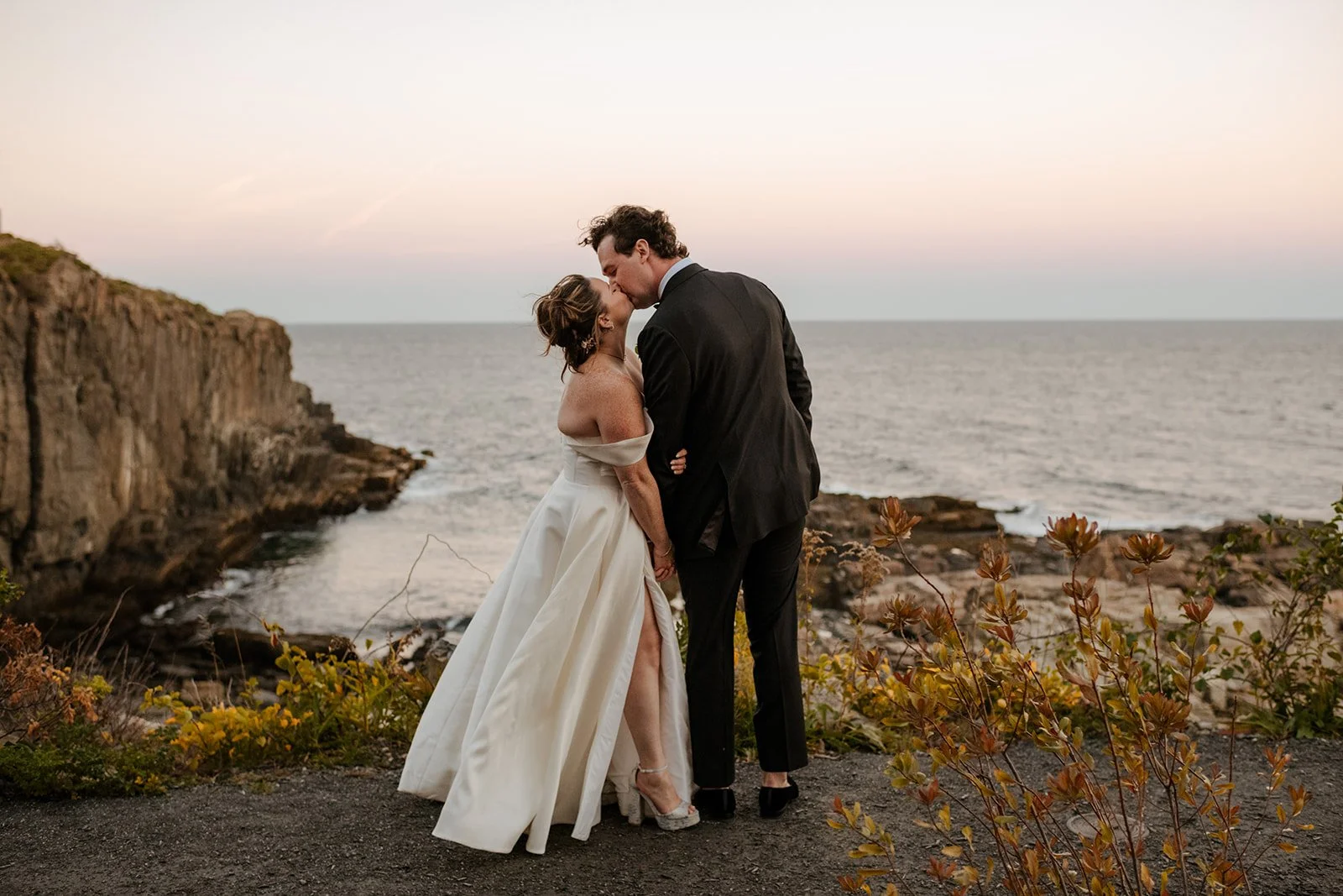 Bride and groom sharing a kiss during sunset portraits on a rocky coastal cliff overlooking the ocean, with soft pastel skies creating a romantic wedding backdrop.