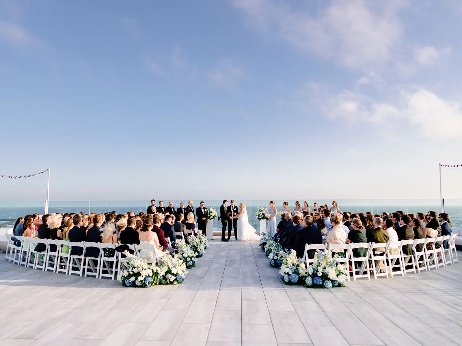 Outdoor oceanfront wedding ceremony on a waterfront terrace, featuring a bride and groom exchanging vows surrounded by guests, white floral arrangements, and ocean views.