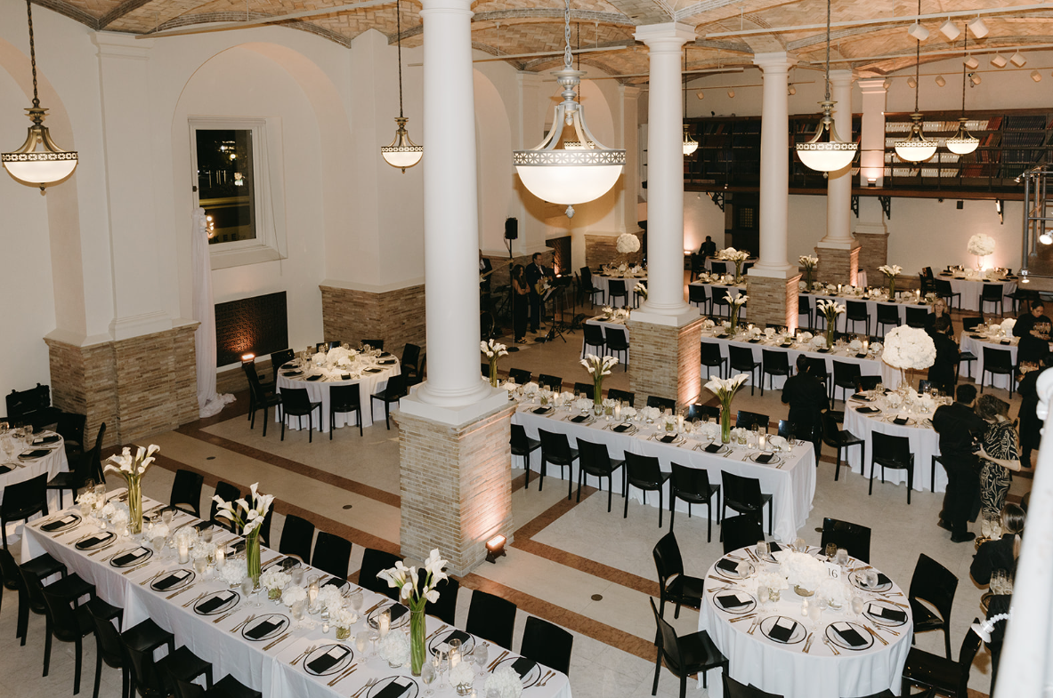 Elegant banquet hall decorated with white tablecloths, tall floral centerpieces, and black chairs. Large chandeliers hang from the ceiling, and soft lighting highlights the space. Several round and long rectangular tables are set up for a formal event.