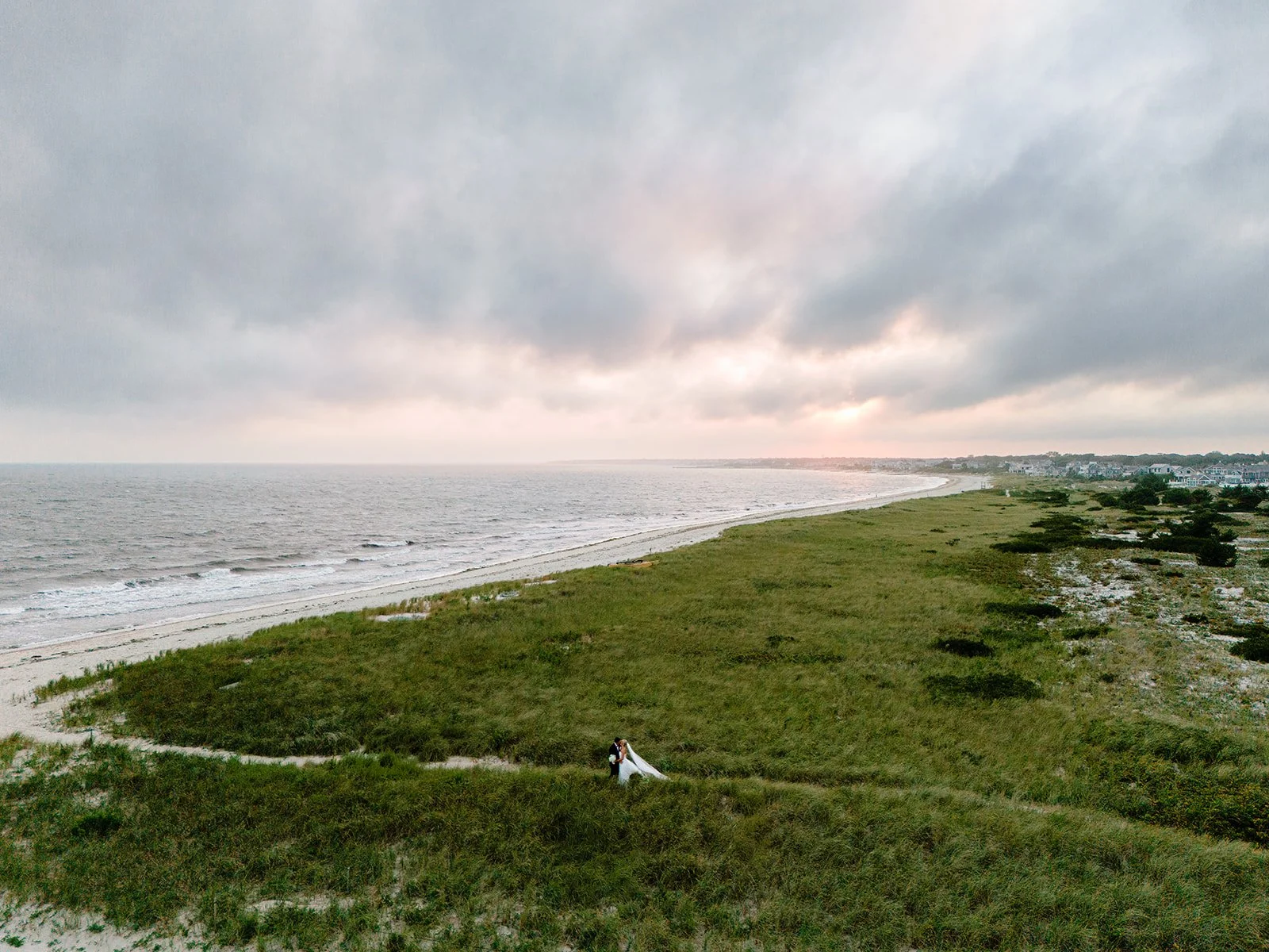 Aerial view of a bride and groom embracing along a scenic coastal shoreline with rolling dunes, the ocean, and a dramatic sunset sky creating a breathtaking wedding portrait.