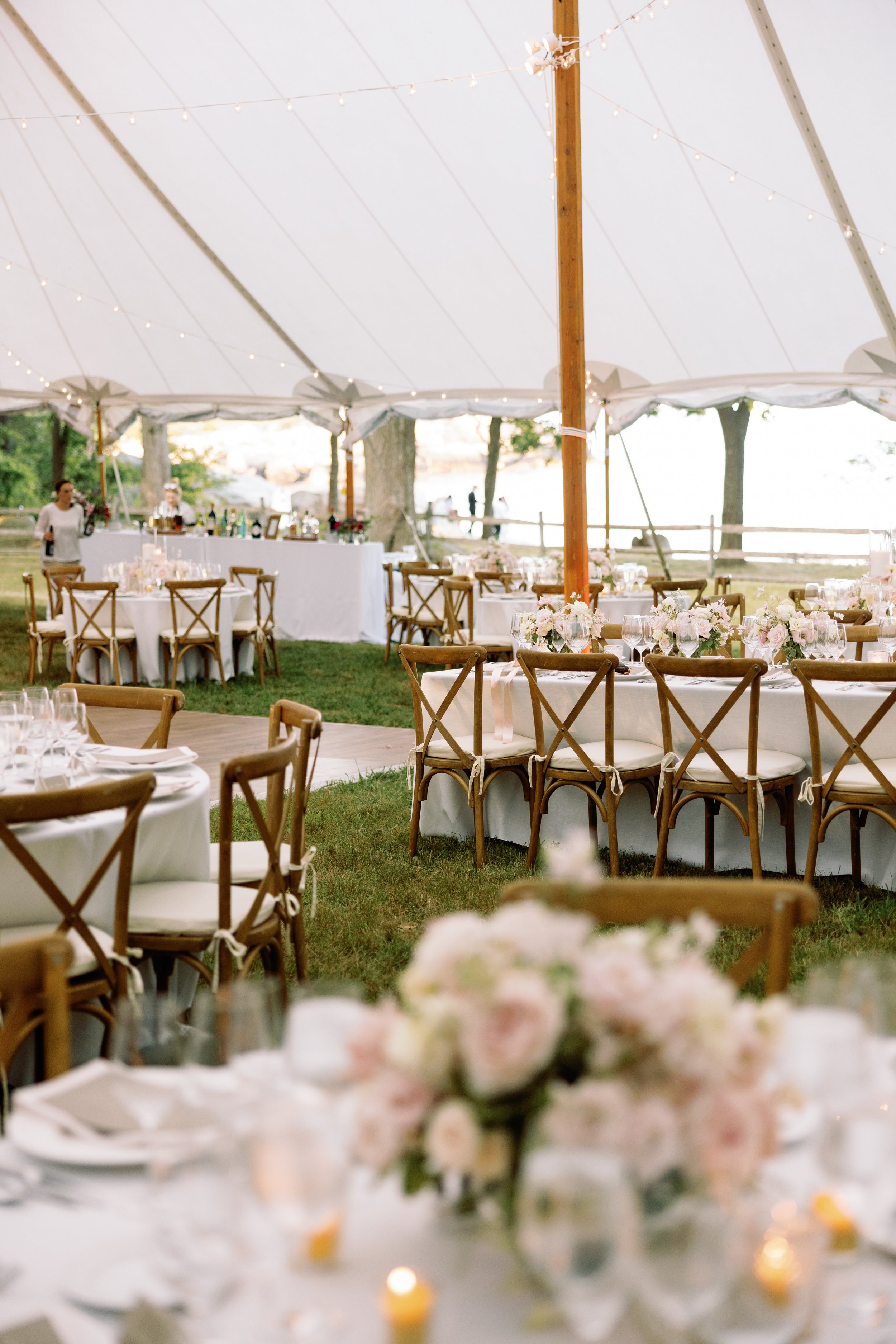 Wedding reception setup under a large white tent with round and rectangular tables decorated with pink and white flowers, glassware, and candles, accompanied by wooden cross-back chairs, outdoor greenery, and string lights hanging from the tent.