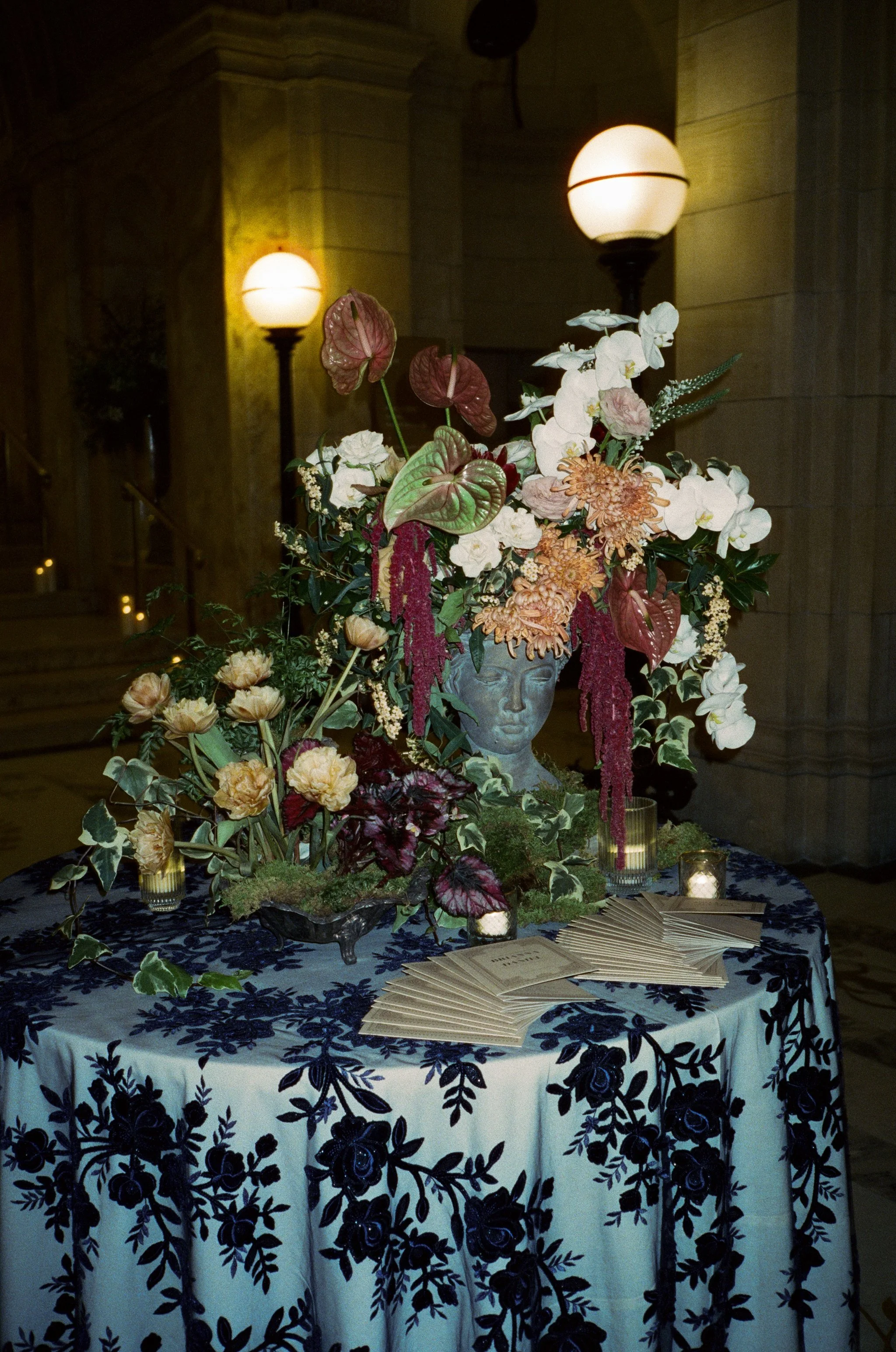 A table with a floral arrangement featuring a face sculpture, candles, and pamphlets in an elegant indoor setting with warm lighting.