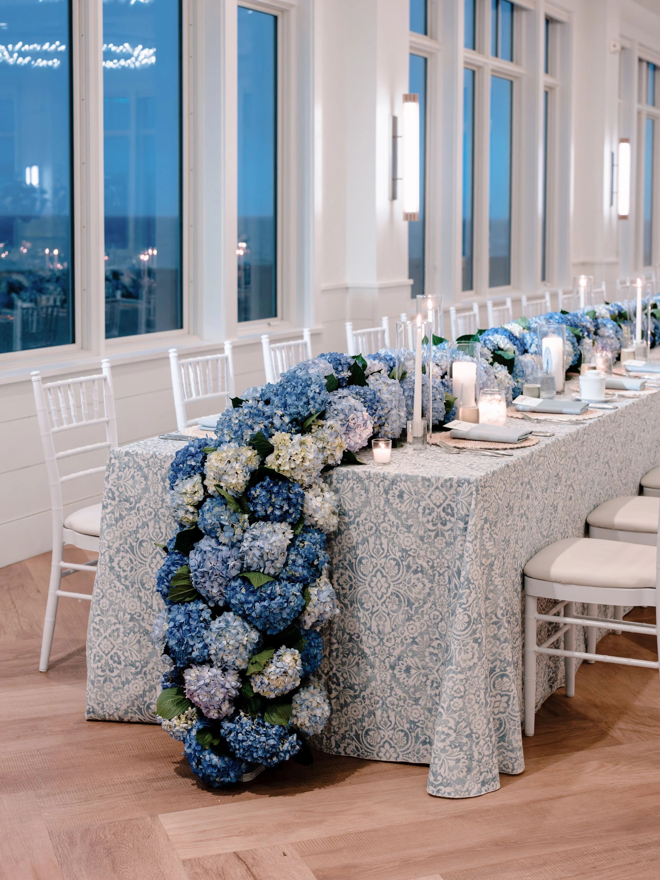 Elegant wedding head table featuring cascading blue and white hydrangeas, candlelight, patterned linens, and ocean-view windows for a timeless coastal reception.