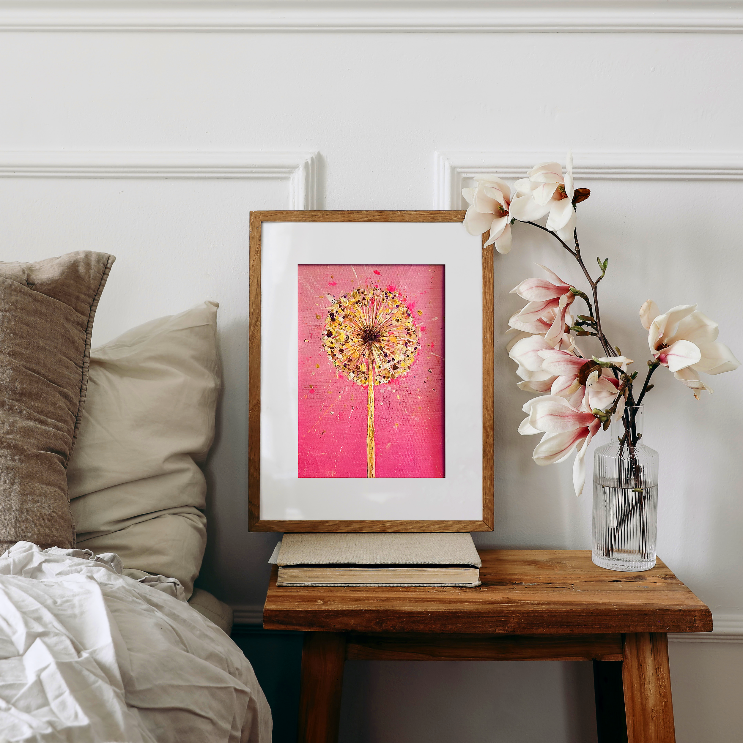 A framed pink artwork of a dandelion on a bedside table next to a white vase with magnolia flowers.