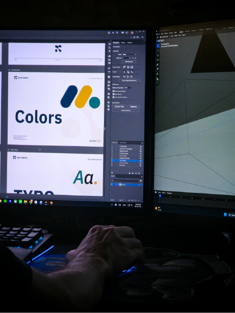 A designer working across two screens: the Sphin Media brand guidelines in Adobe Illustrator, a 3D scene in Blender.