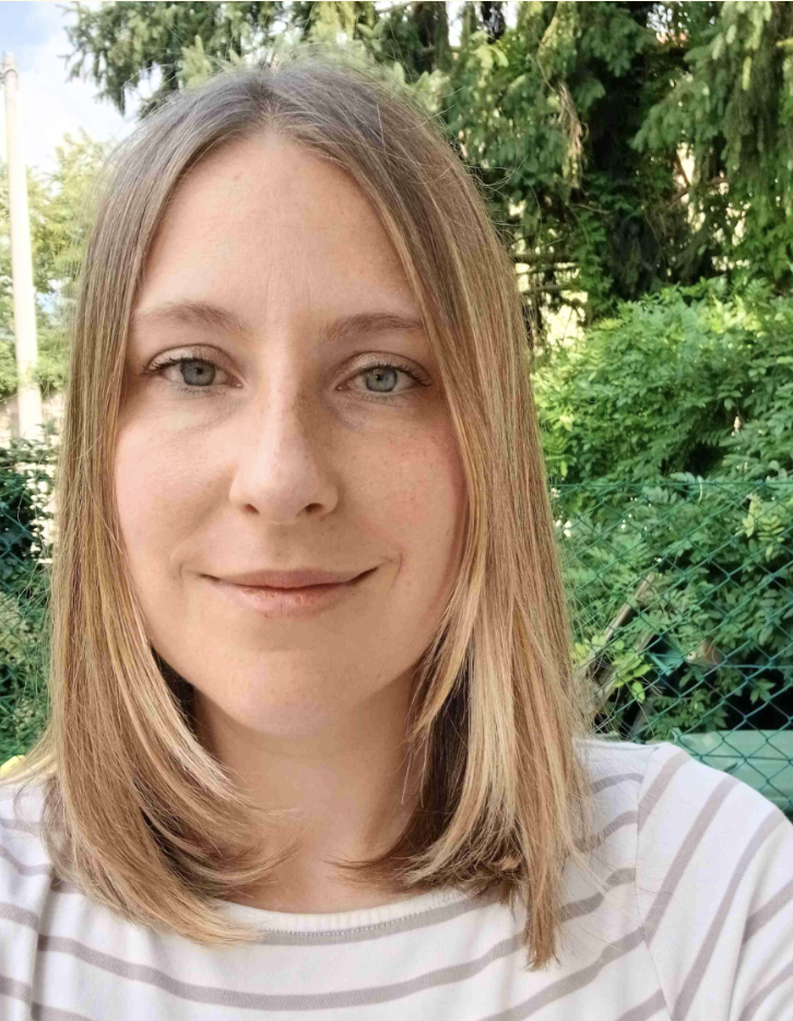 Frau mit langen, welligen braunen Haaren und einem weißen Oberteil vor einem neutralen Hintergrund