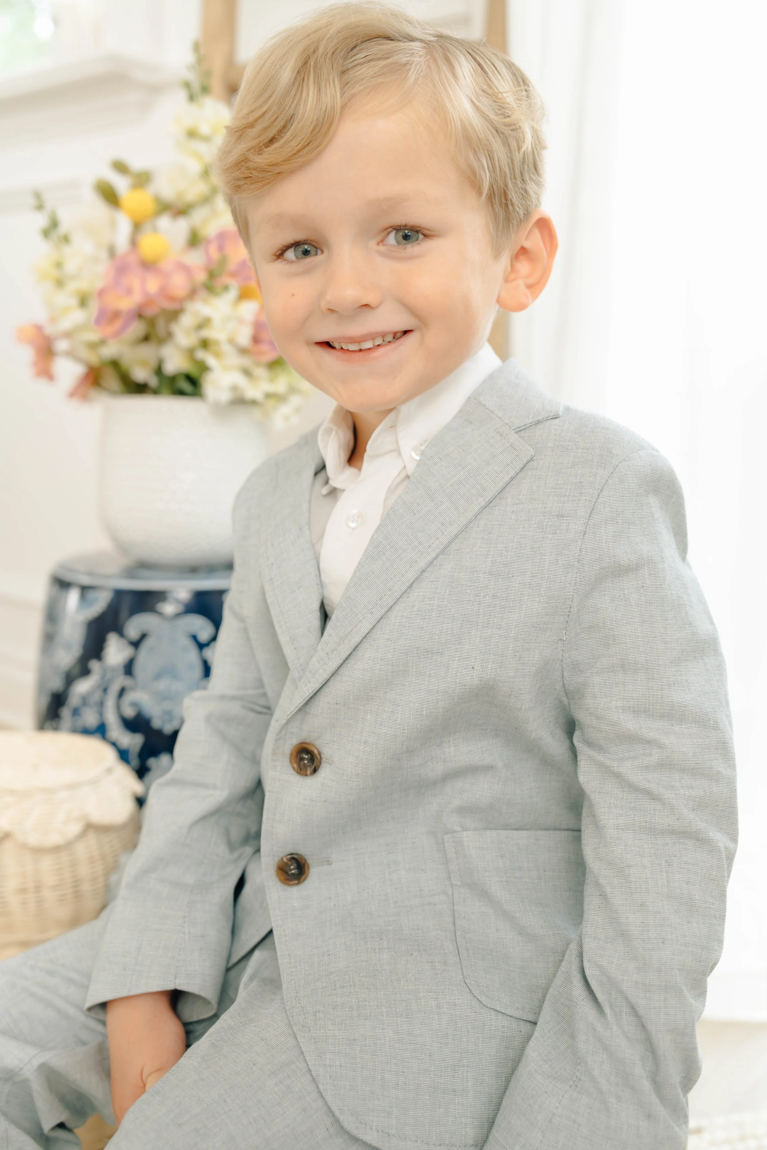 A young boy with blonde hair and blue eyes wearing a light grey blazer and white shirt, smiling at the camera, with a vase of colorful flowers in the background.