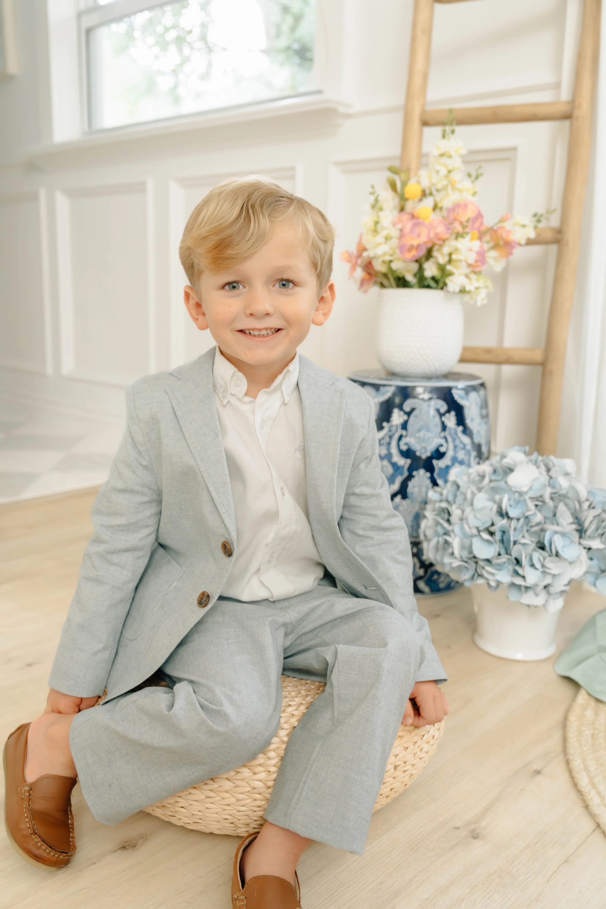A young boy with blonde hair, wearing a light gray suit and brown shoes, sitting on a wicker ottoman in a bright, decorated room with flowers and a ladder.