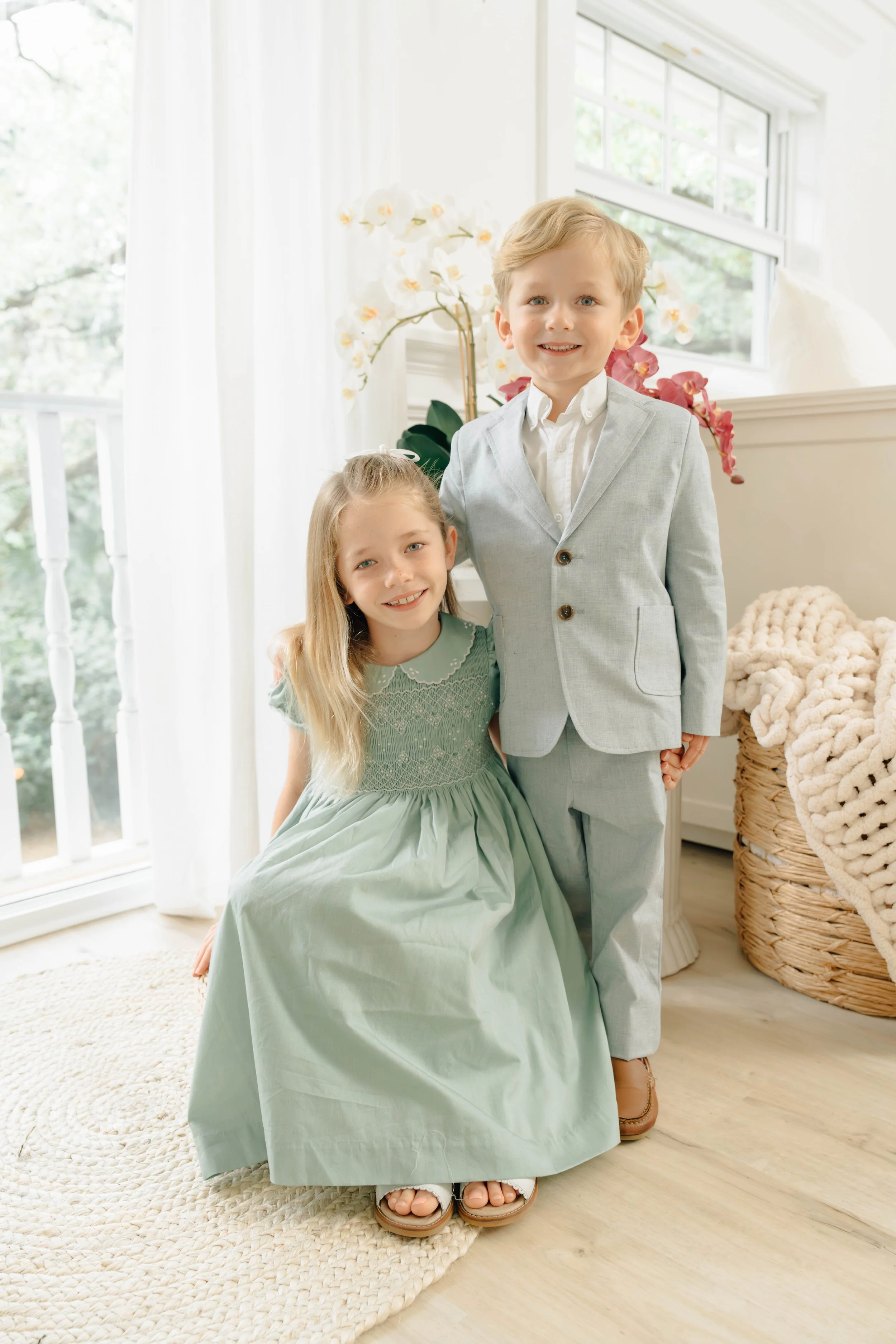 A boy and a girl standing indoors near a window, dressed in formal attire. The girl is sitting on a rug, wearing a light green dress and sandals, while the boy is standing beside her in a light gray suit with brown shoes. Behind them are white curtai