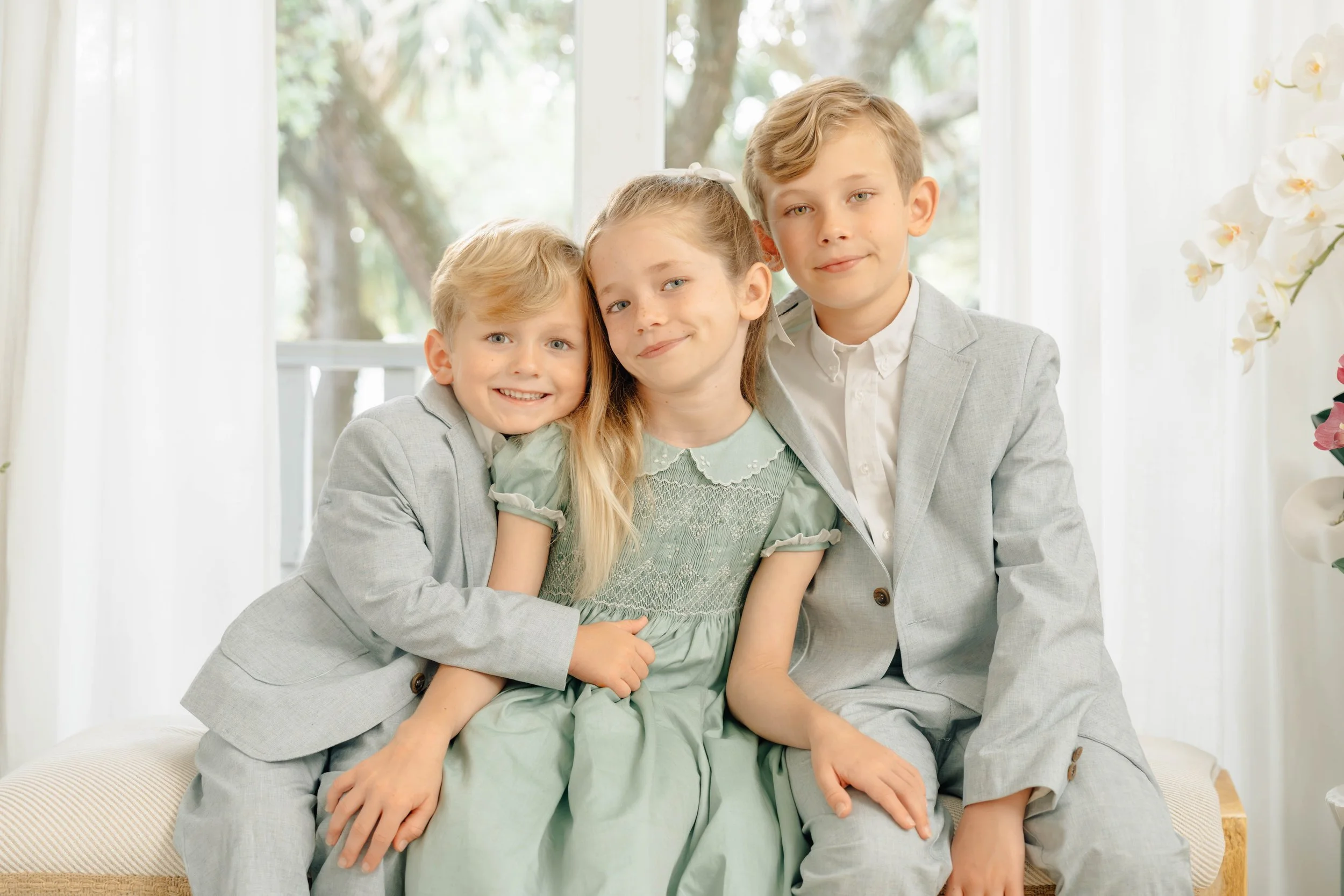 Three children sitting on a beige cushion in front of a large window with white curtains and trees outside. The children are dressed in light-colored formal clothing, with the girl in the middle wearing a light green dress and the two boys on either 