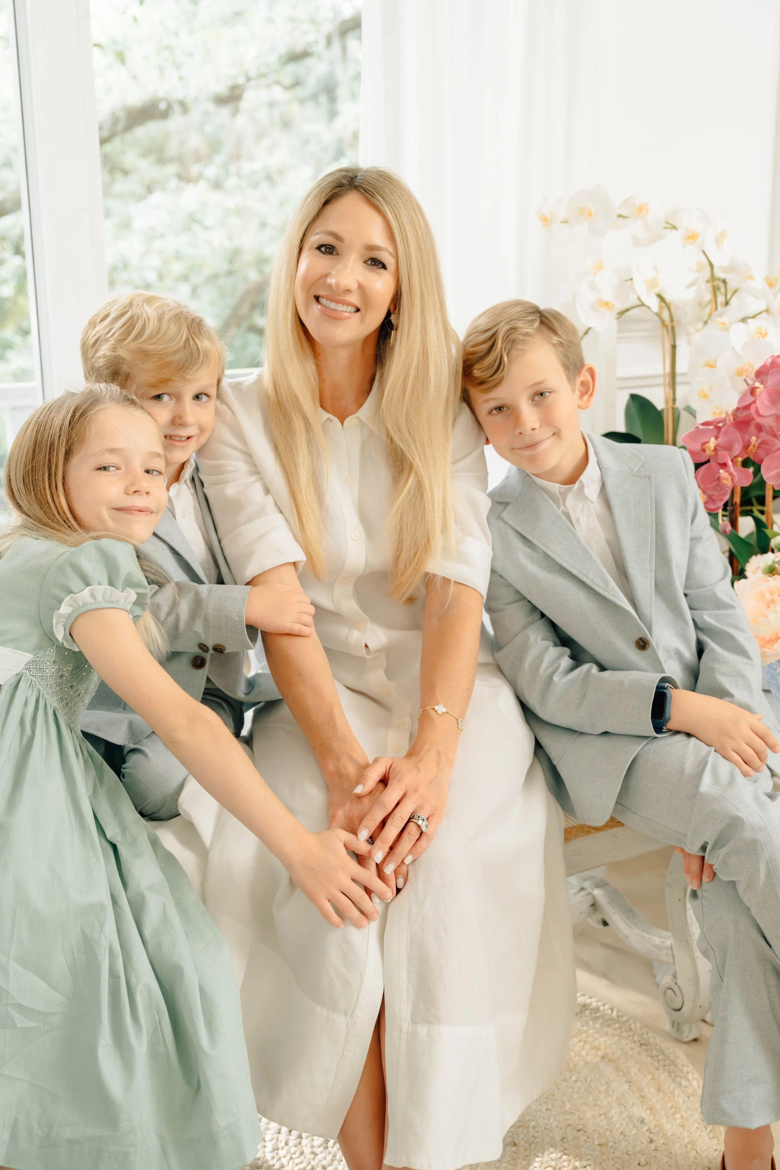 A woman sitting with four children, all dressed in light-colored formal clothes, in a bright room with large windows and white flowers in the background.