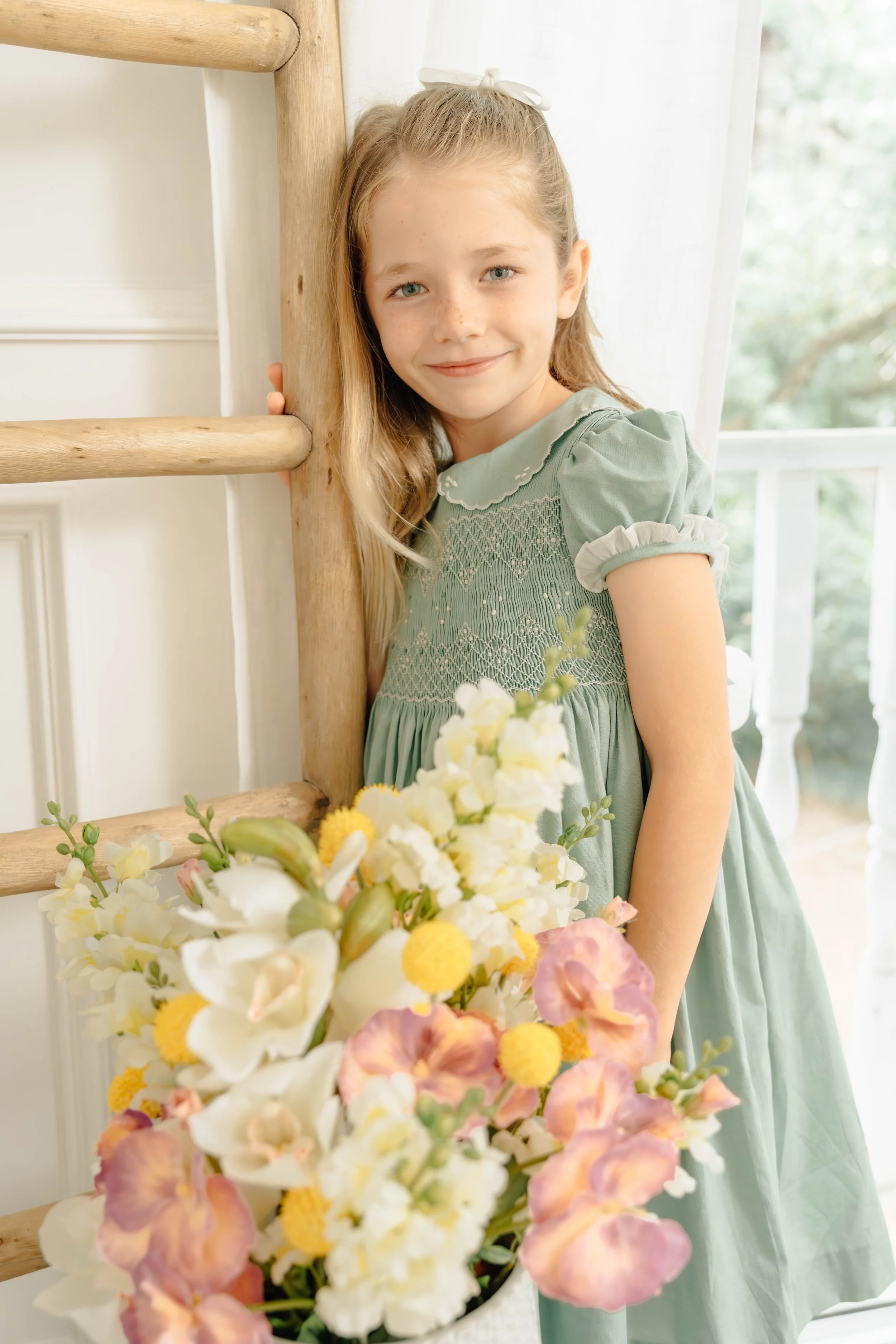 A young girl with long blonde hair, wearing a light green dress with puffed sleeves, standing next to a wooden ladder, holding a large bouquet of white, pink, and yellow flowers, indoors near a window with white curtains.