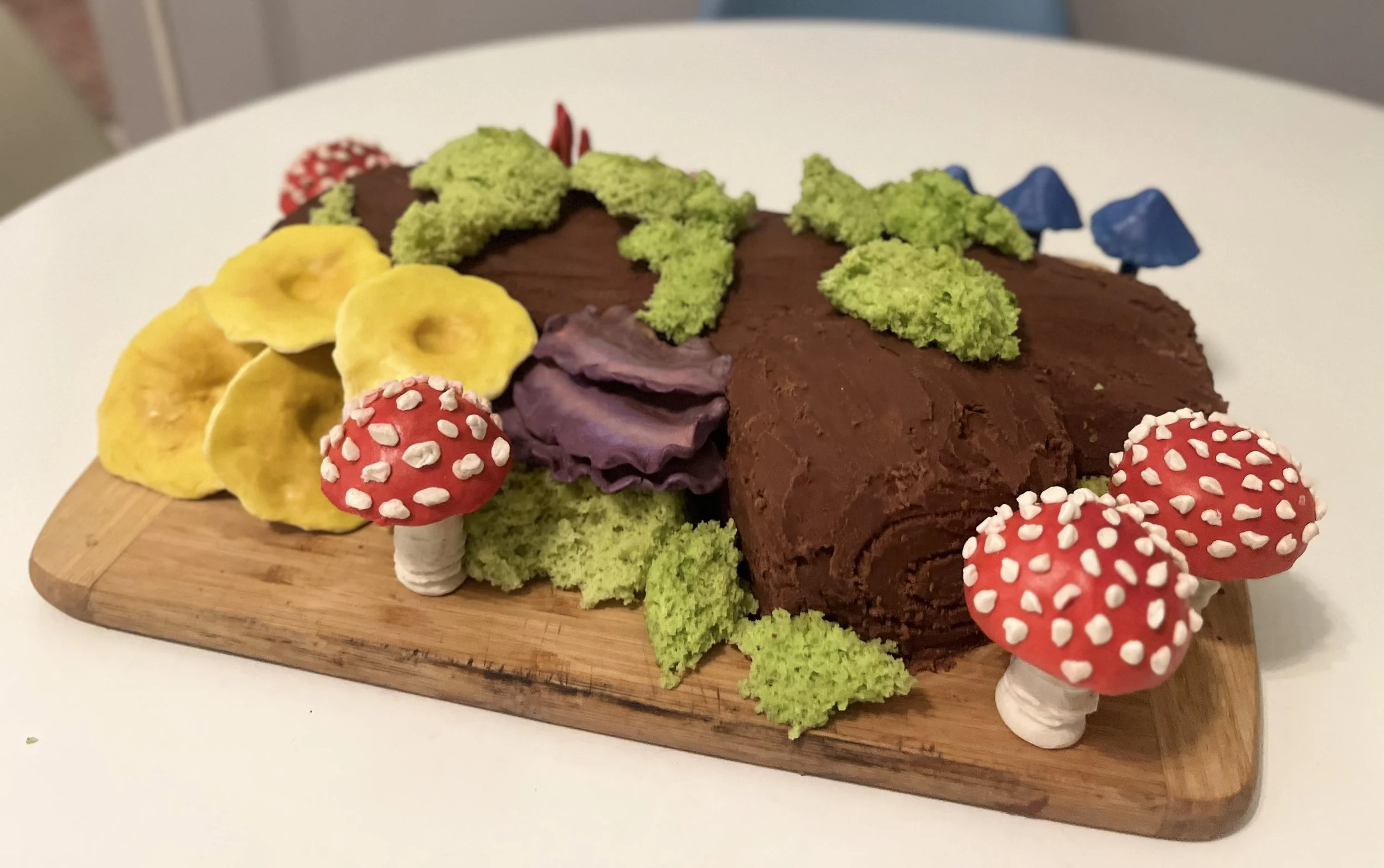 A miniature chocolate cake designed to look like a forest scene with colorful candy mushrooms, green sponge trees, yellow banana chips, and blue rock-like candies on a wooden board.
