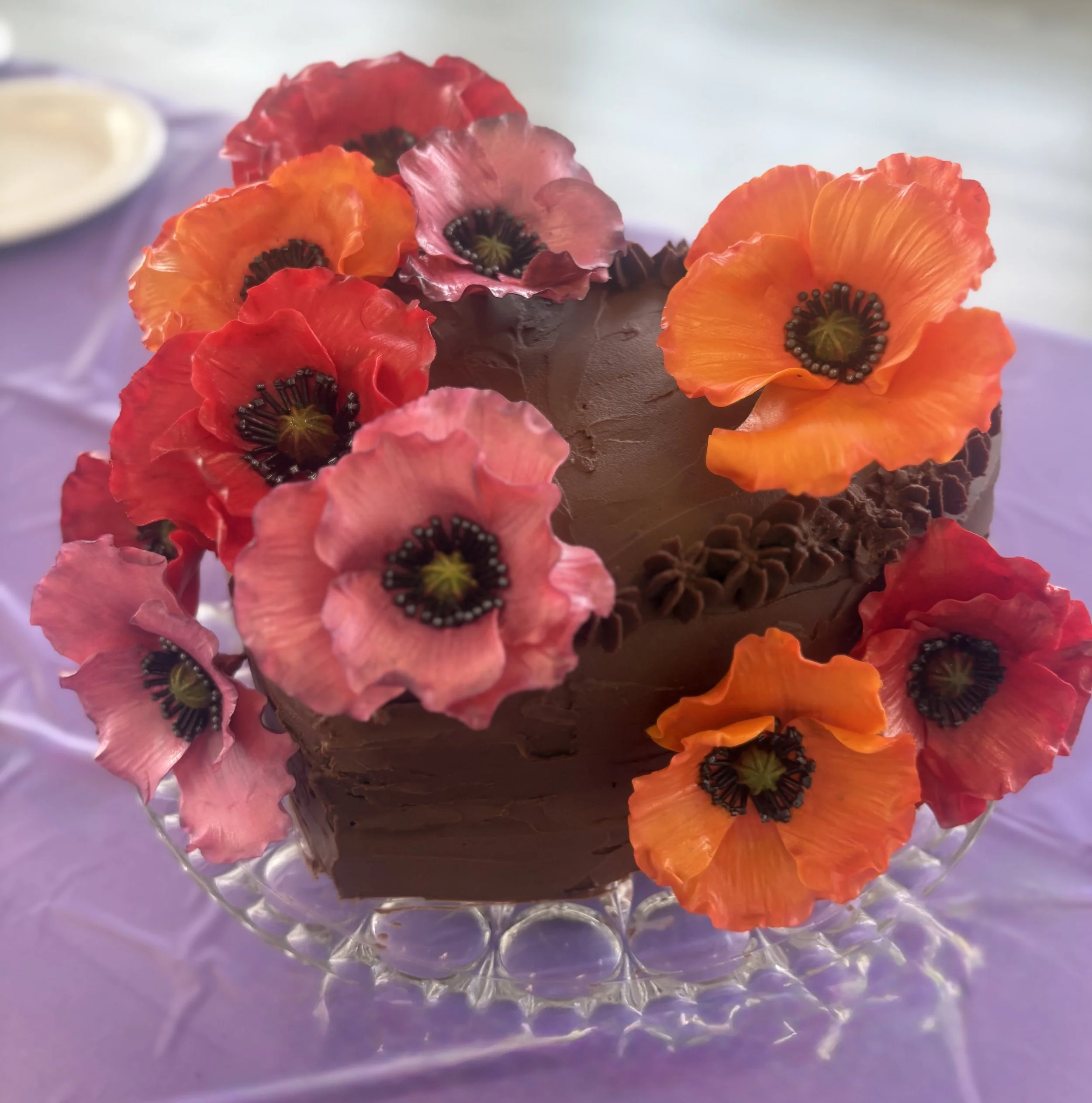 Chocolate cake decorated with colorful edible flowers on top, placed on a glass cake stand.