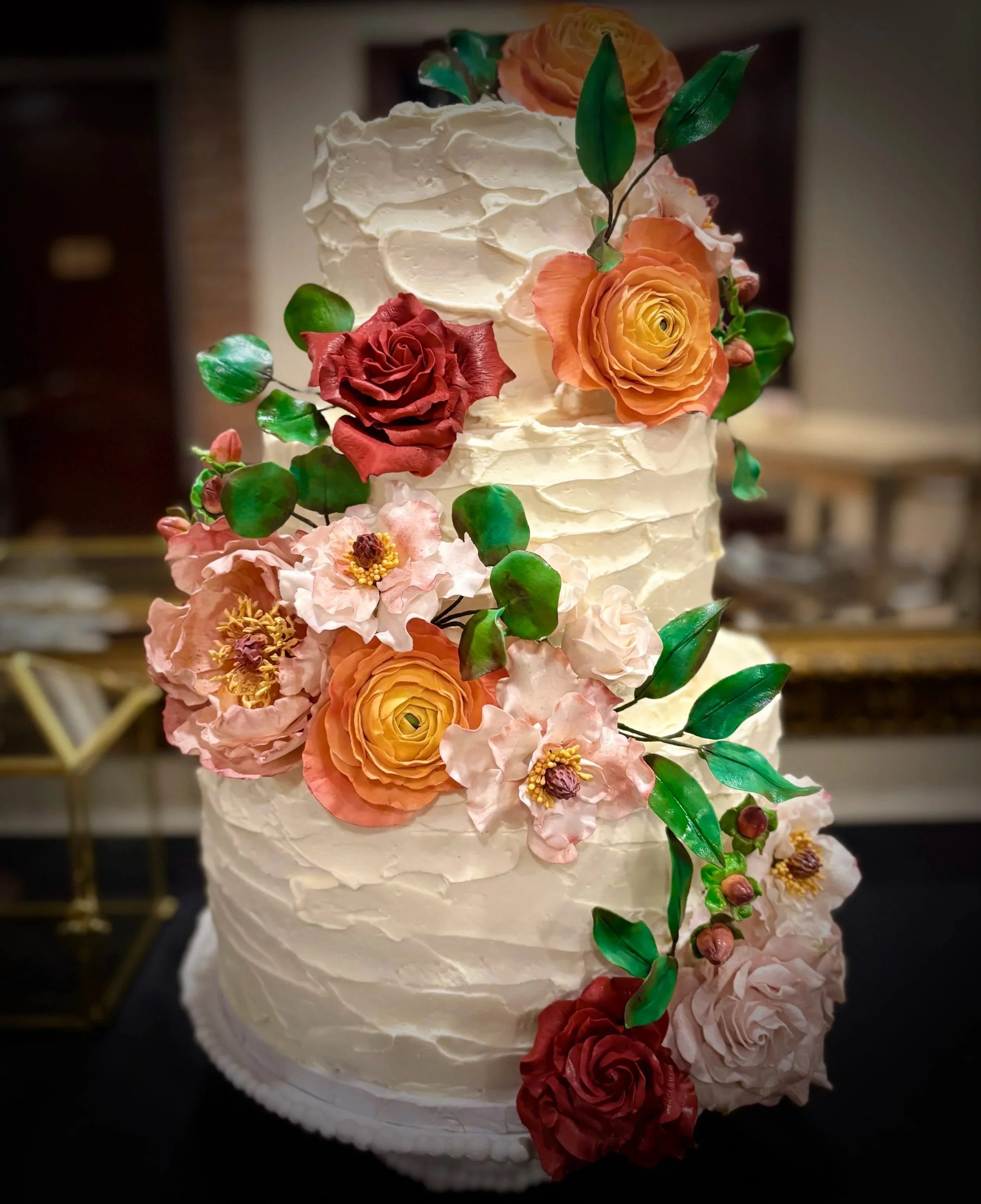 Three-tier wedding cake decorated with red, pink, and peach sugar flowers