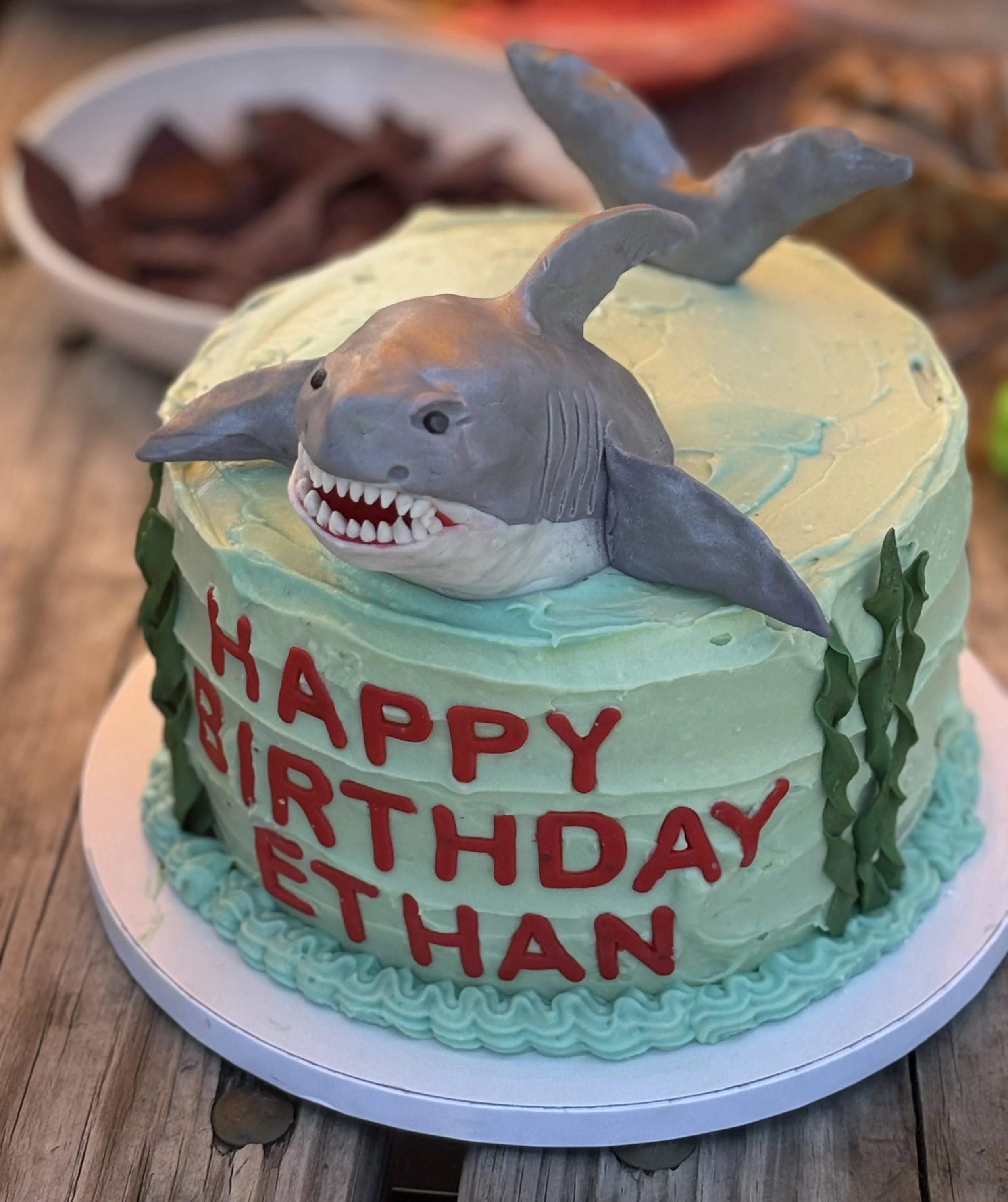 A birthday cake with a shark figure on top, decorated with green and blue icing, and red letters saying "Happy Birthday Ethan".