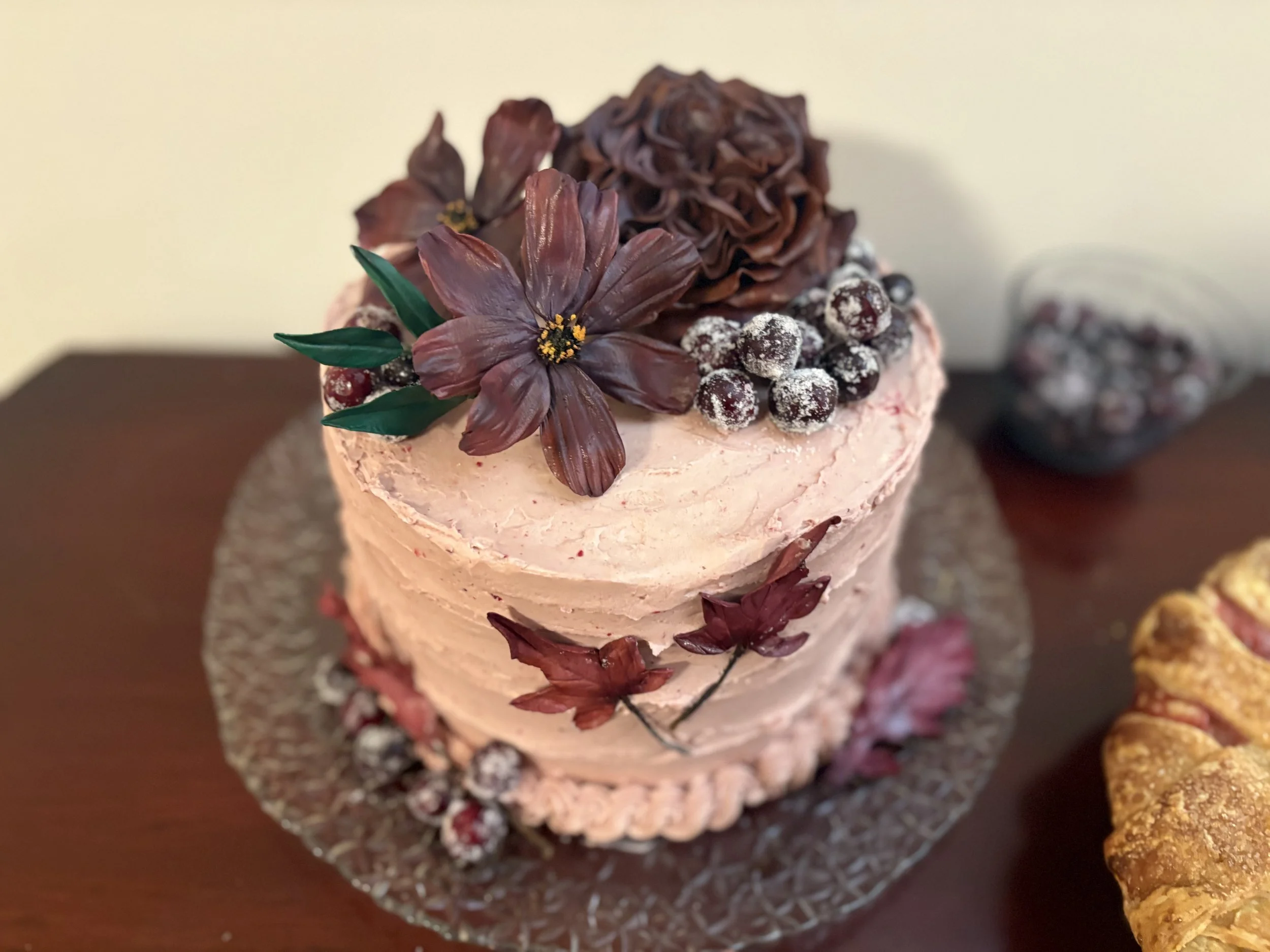 A pink-frosted cake decorated with edible flowers, berries, and leaves, placed on a glass plate on a wooden surface.