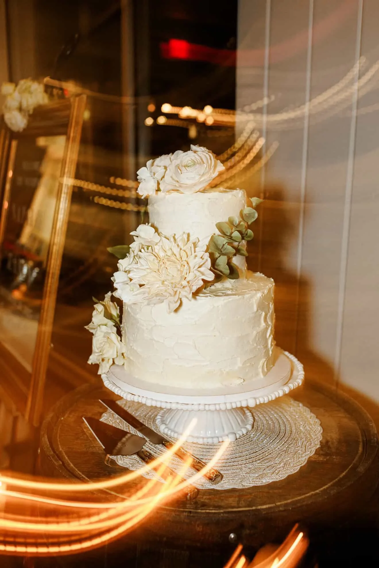 A three-tiered wedding cake with white frosting and floral decorations, displayed behind glass.