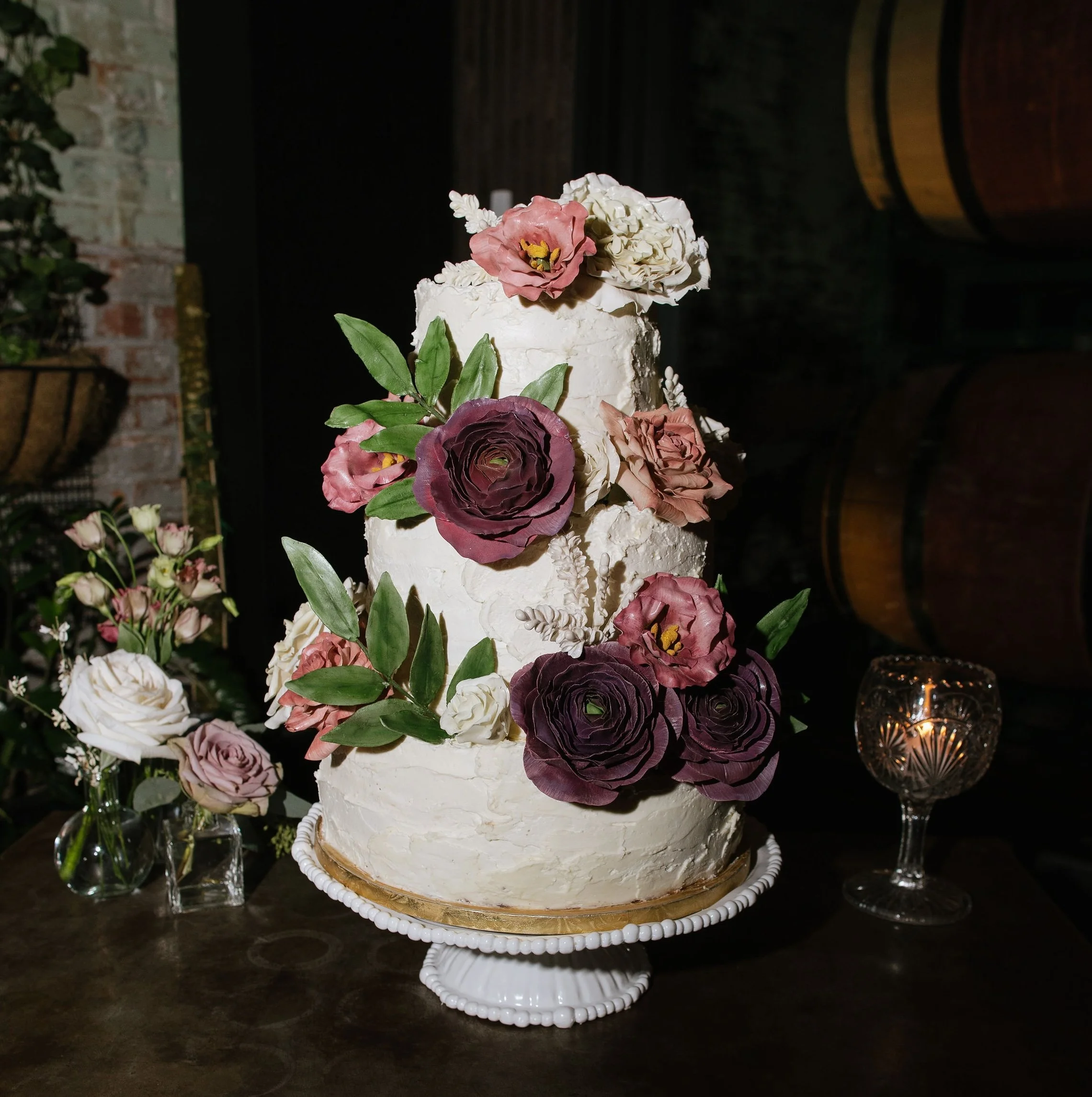 Three-tiered white wedding cake decorated with pink, white, and purple sugar flowers and green leaves, displayed on a white stand.