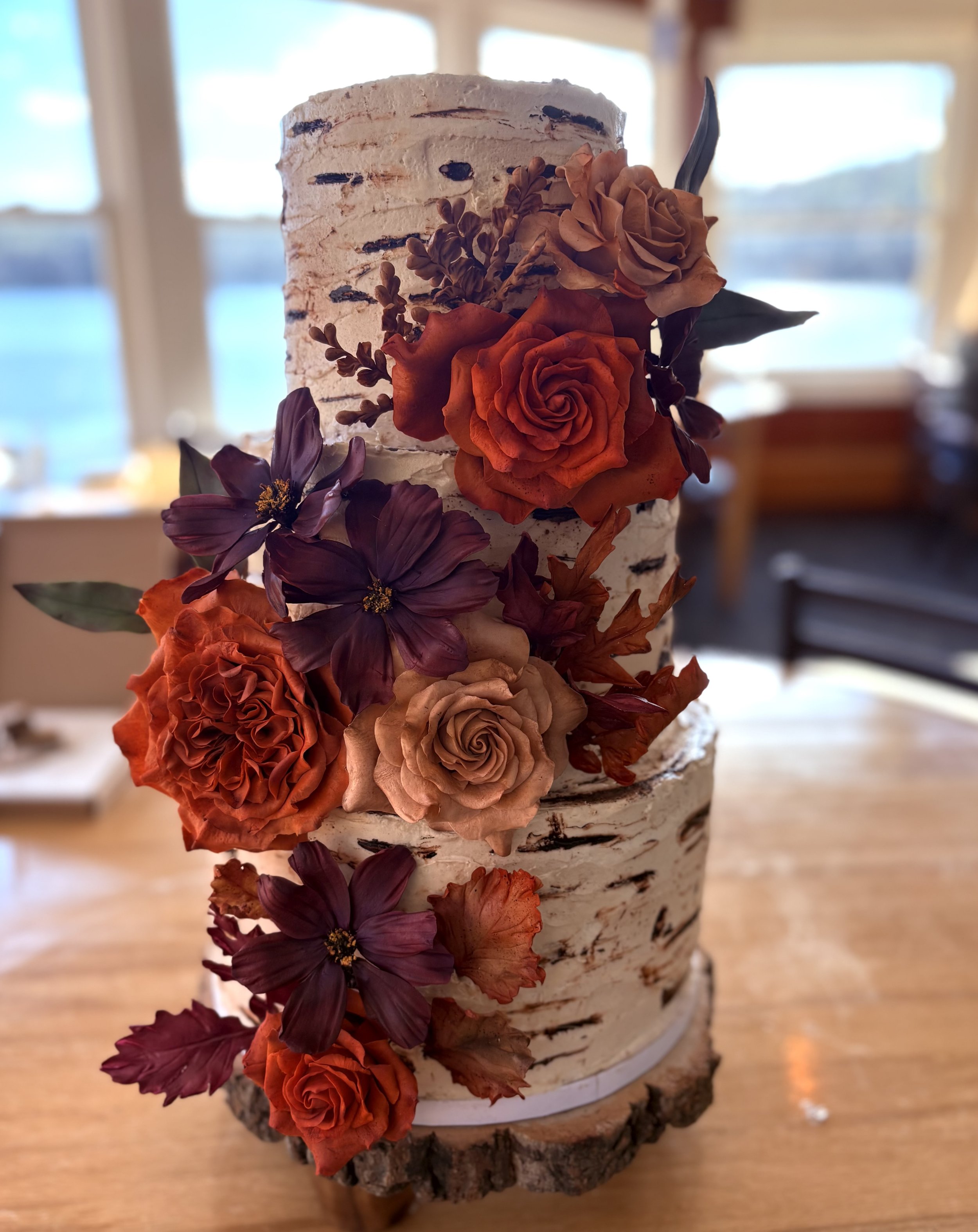A two-tiered cake designed to look like birch wood, decorated with orange, purple, and beige sugar flowers, sitting on a wooden base.