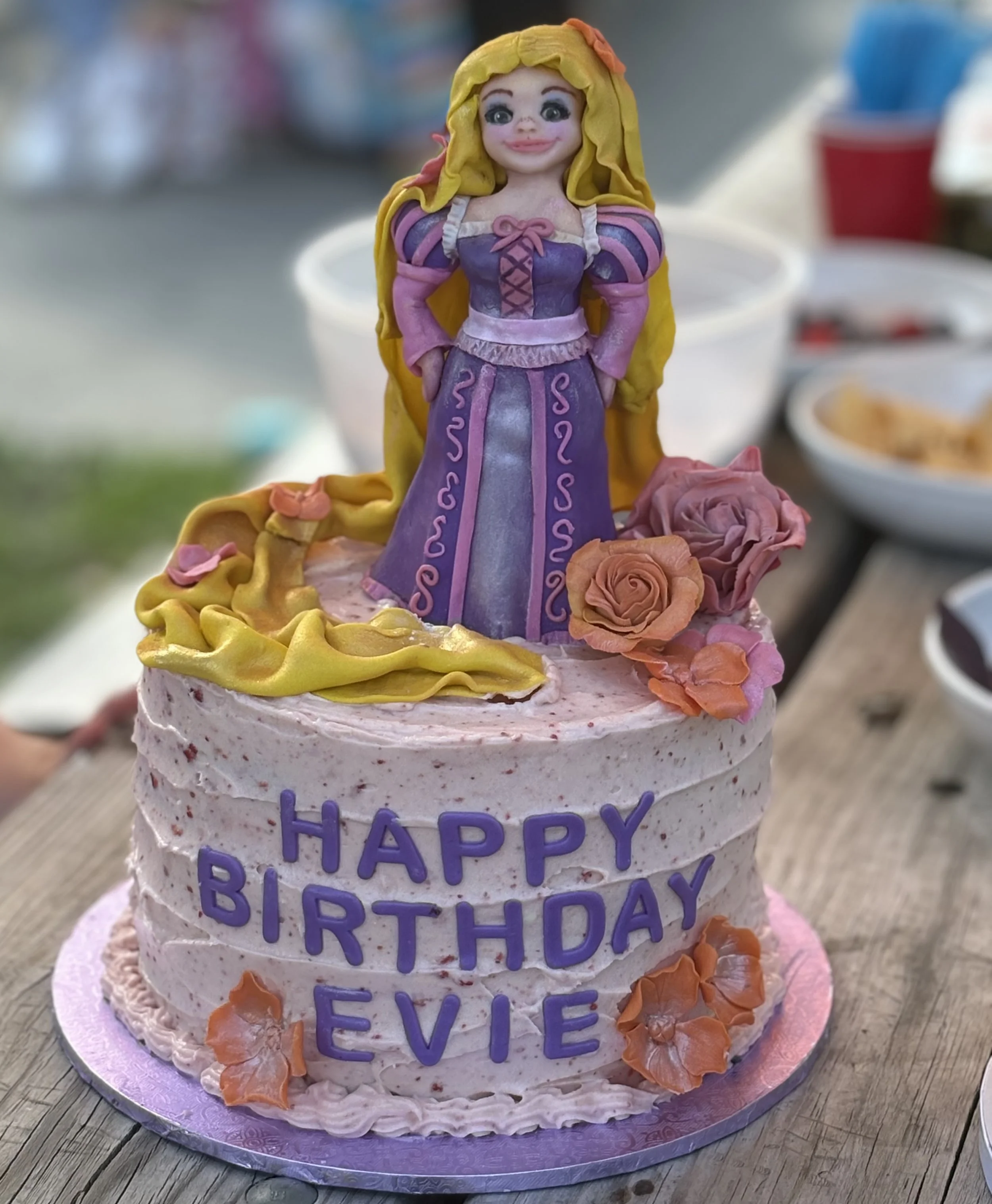 A birthday cake decorated with a princess figurine on top, pink and purple flowers, and the message 'Happy Birthday Evie' in purple letters.