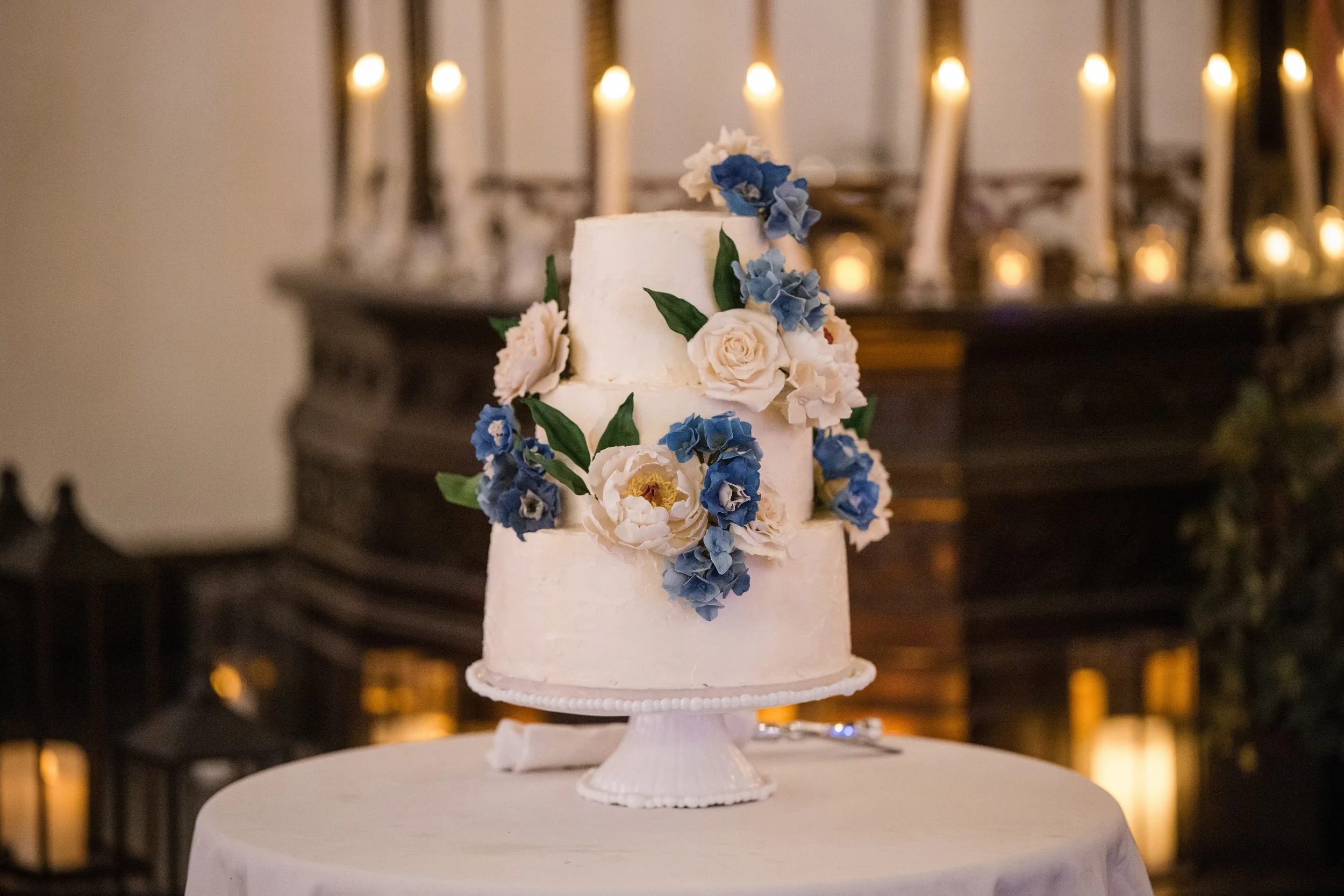A two-tiered white wedding cake decorated with white and blue sugar flowers, displayed on a white cake stand on a round table with a white tablecloth, with candles and a mirror in the background.
