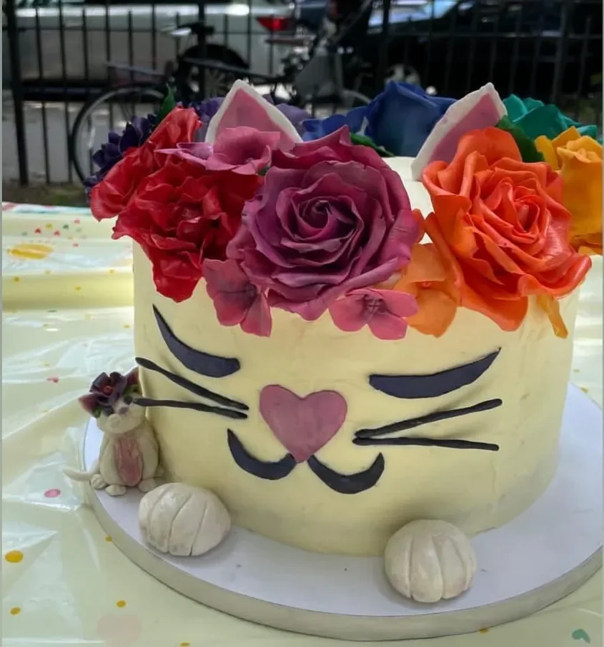 Cake decorated with colorful roses on top and a cat face with closed eyes, whiskers, and a heart-shaped pink nose, along with a small fondant bunny and pumpkin decorations.