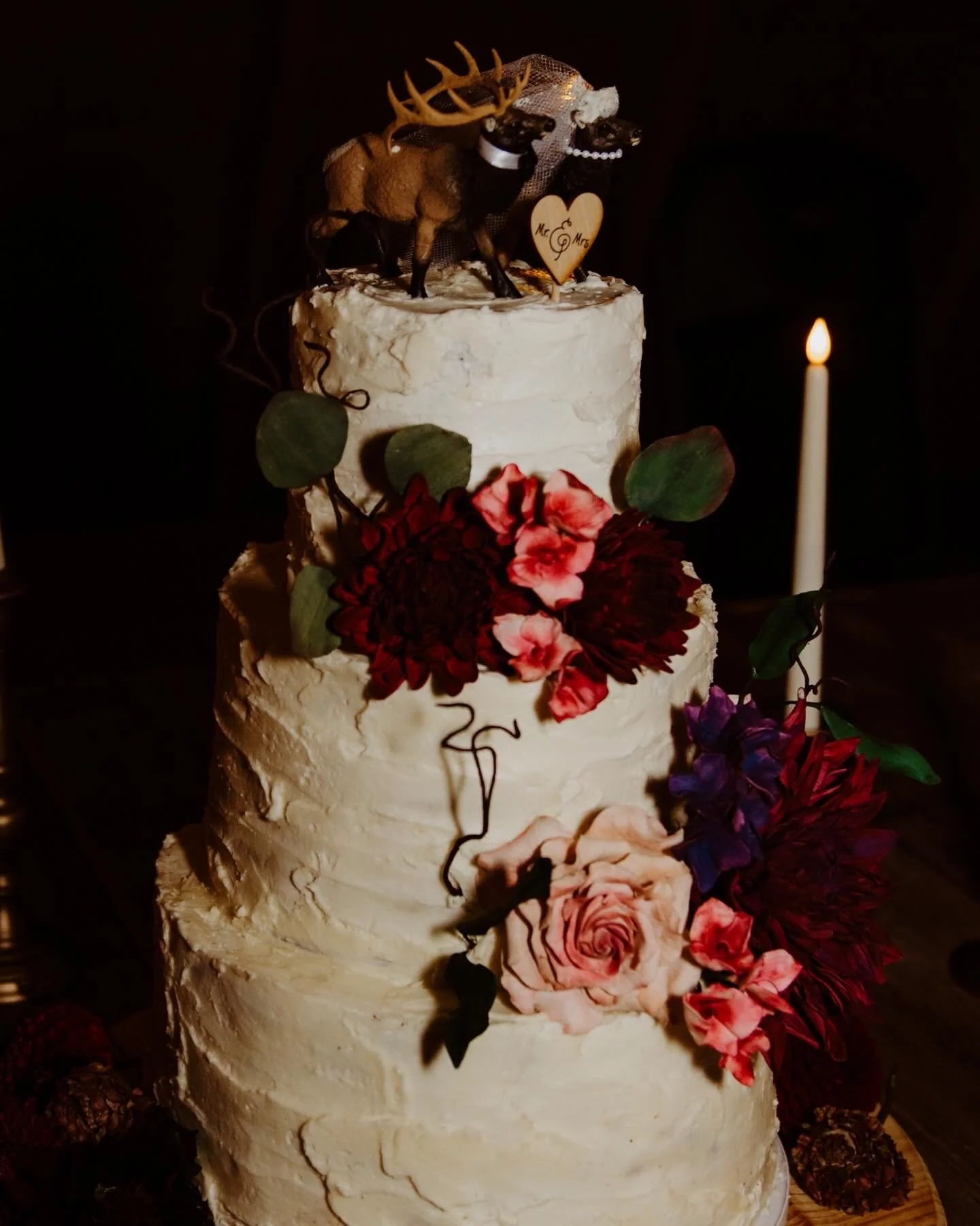 Three-tier white wedding cake decorated with pink, red, and purple flowers, green leaves, and a moose figurine on top with a 'Mr. & Mrs.' heart. A single lit candle is beside the cake.