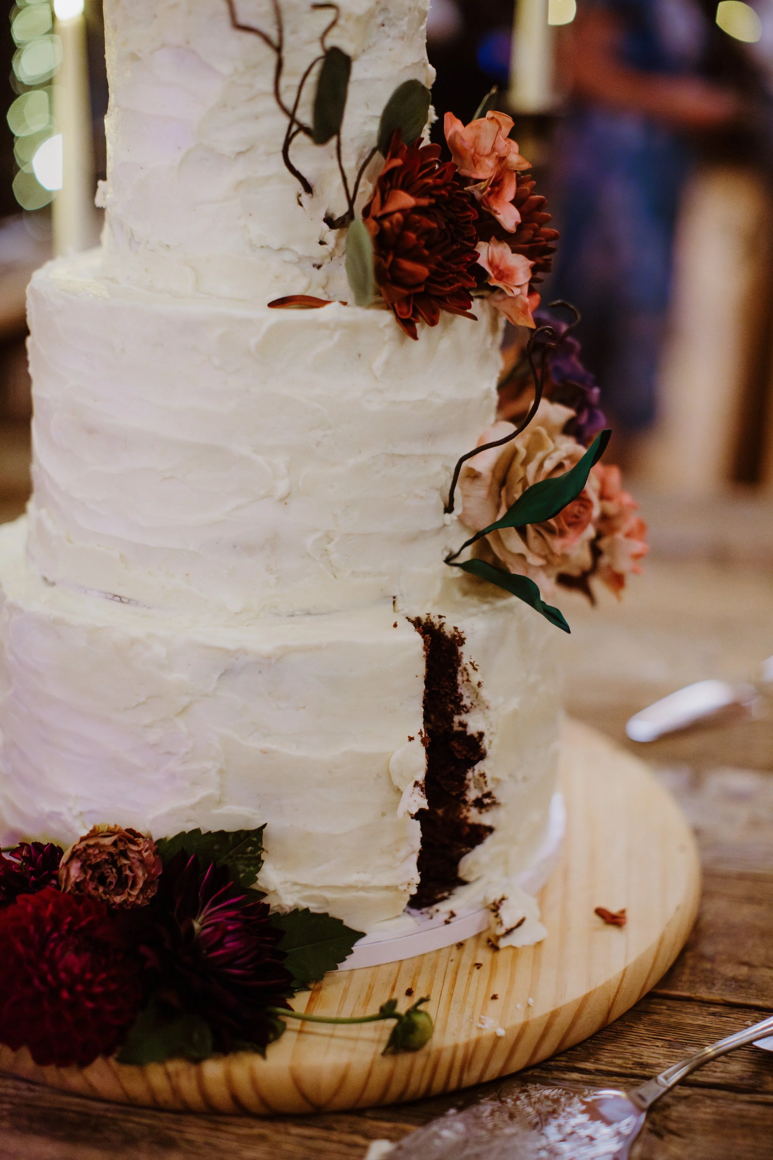 A three-tiered wedding cake with white frosting and floral decorations, featuring blush, burgundy, and mauve flowers and green leaves, on a wooden cake board with some crumbs and flowers around it.