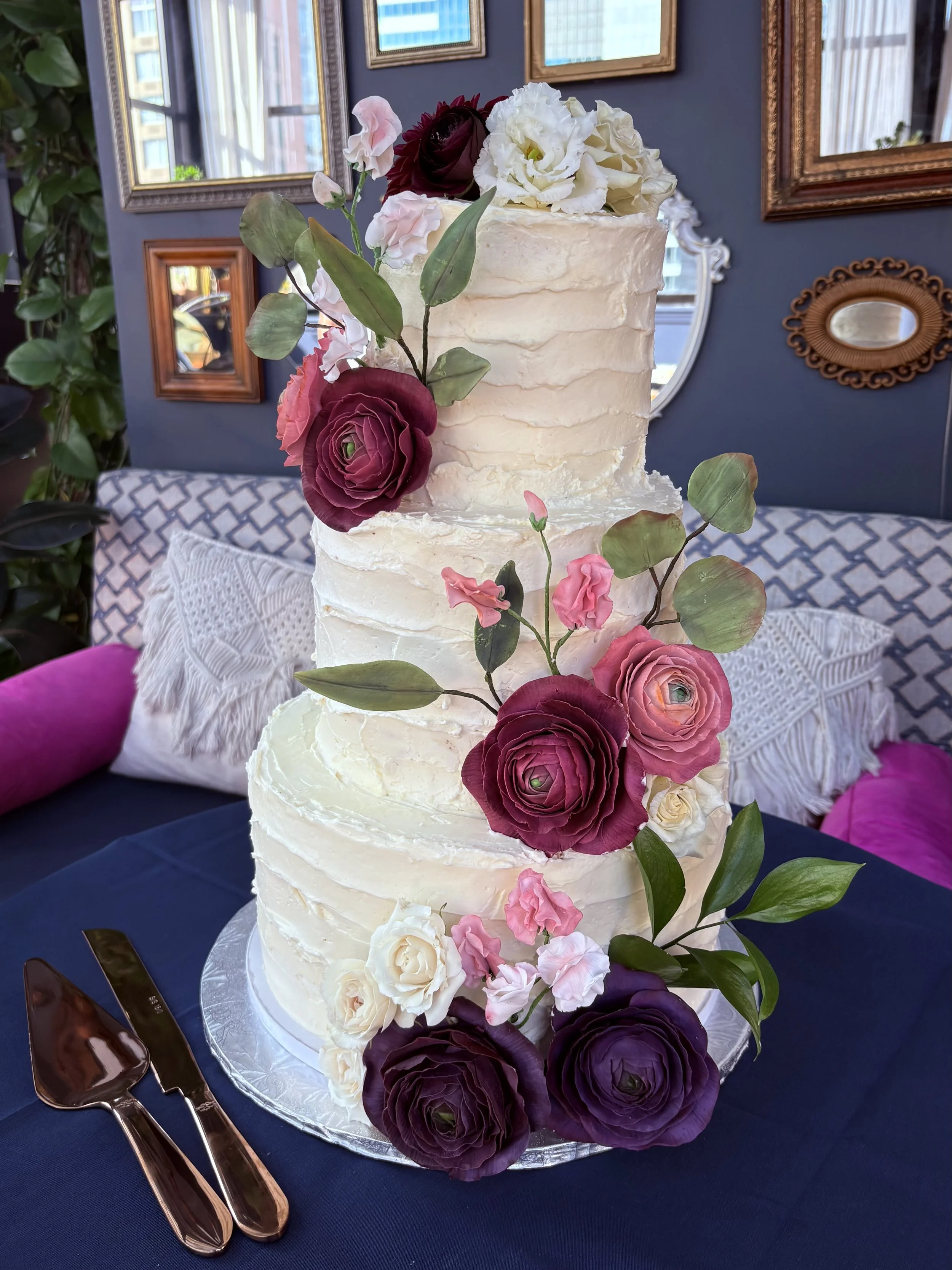Three-tier wedding cake decorated with white frosting and pink, purple, and white sugar flowers, placed on a table with a cake server and knife in front of it.