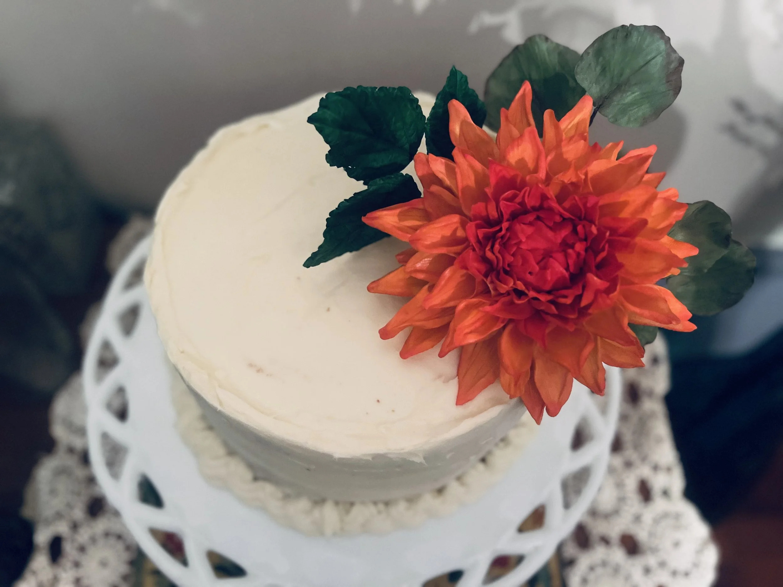 A round white frosted cake decorated with a large orange and red artificial flower and green leaves, placed on a white doily cake stand.