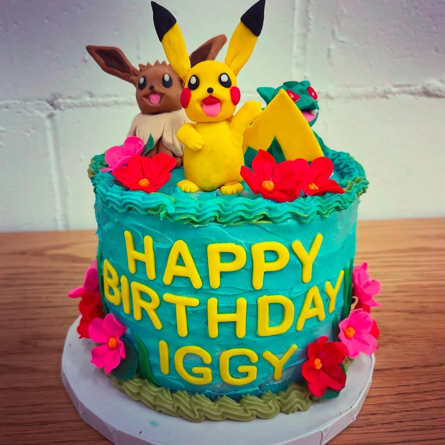 A colorful birthday cake decorated with Pokémon characters Pikachu, Eevee, and Bulbasaur, with flowers and the words 'Happy Birthday Iggy' written on it.