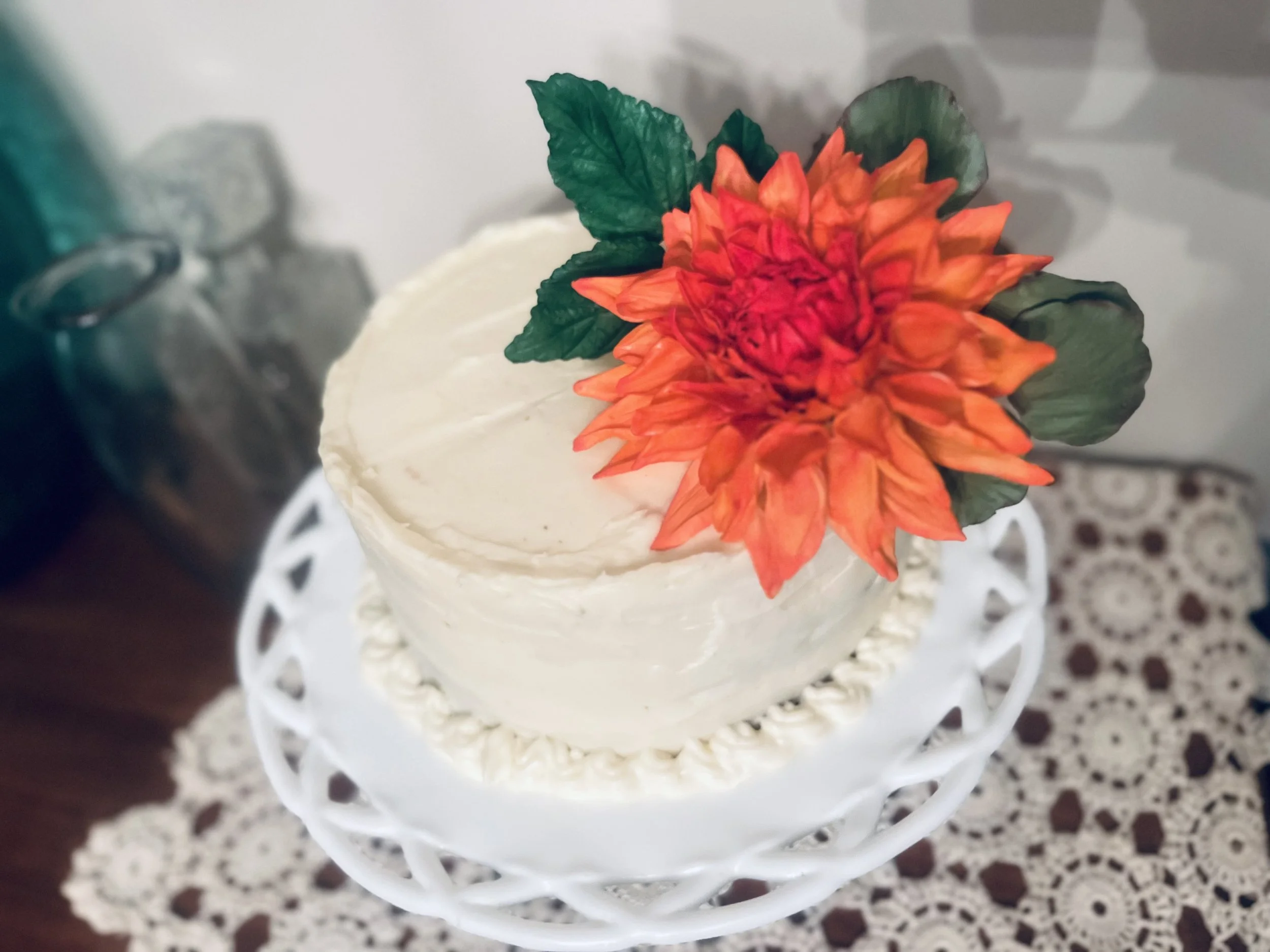 A small, frosted cake with a large orange and red flower and green leaves on top, served on a white decorative cake stand.