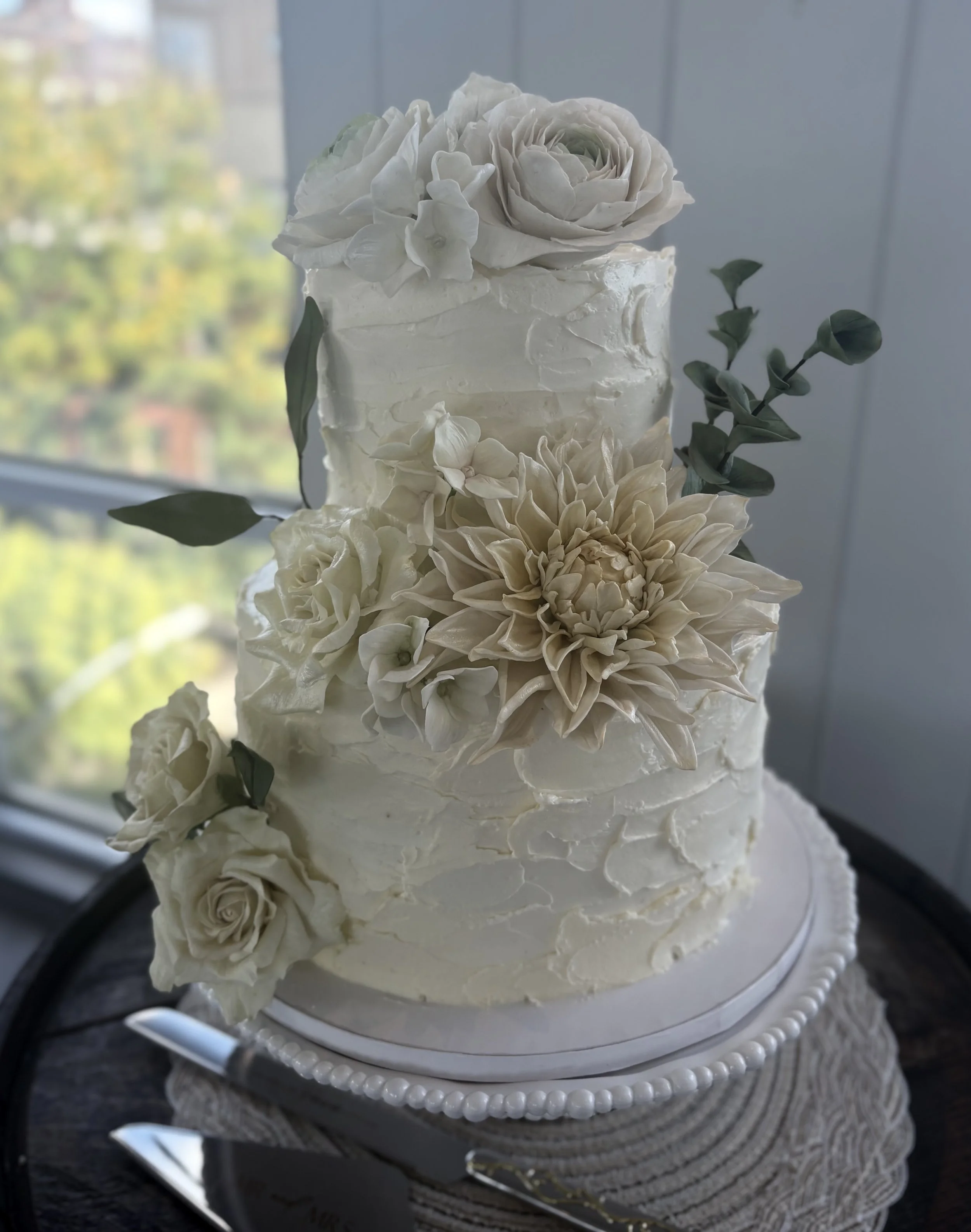Elegant three-tier wedding cake decorated with white sugar flowers and greenery, set on a decorated cake stand near a window.