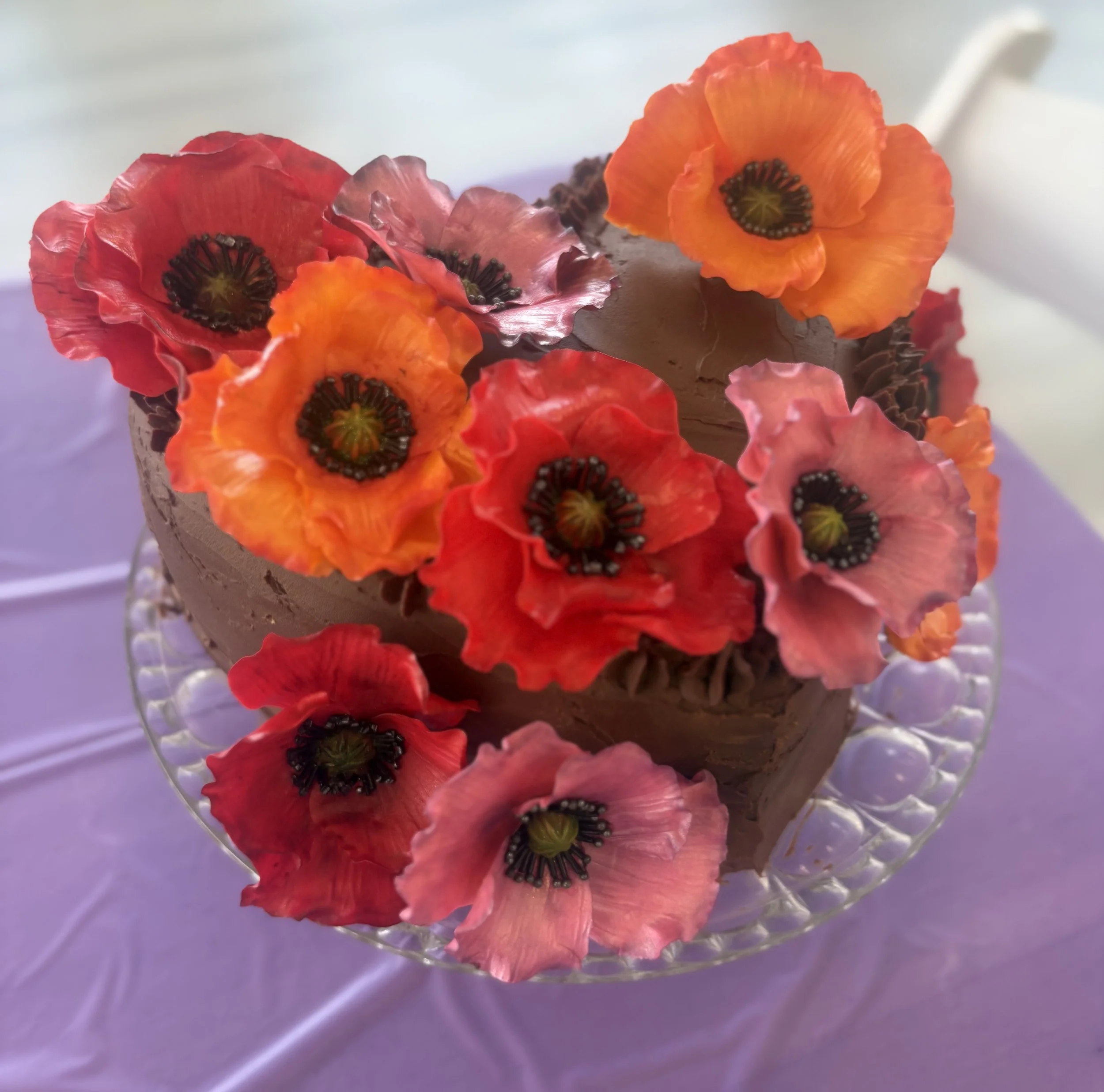 Chocolate cake decorated with colorful edible poppy flowers on a glass cake stand.
