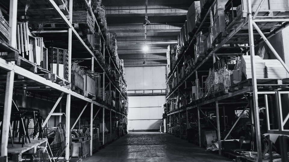 A large warehouse with tall metal shelves stocked with various items such as chairs, boxes, and supplies, illuminated by industrial lighting.