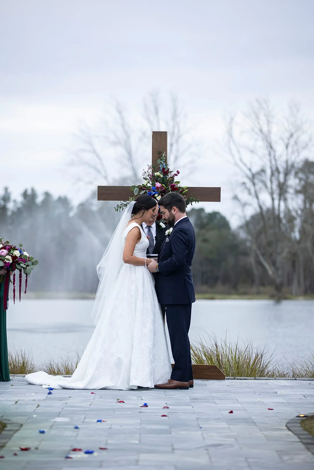 Bride and groom holding hands during outdoor wedding ceremony in front of a wooden cross with floral decorations, near a lake.