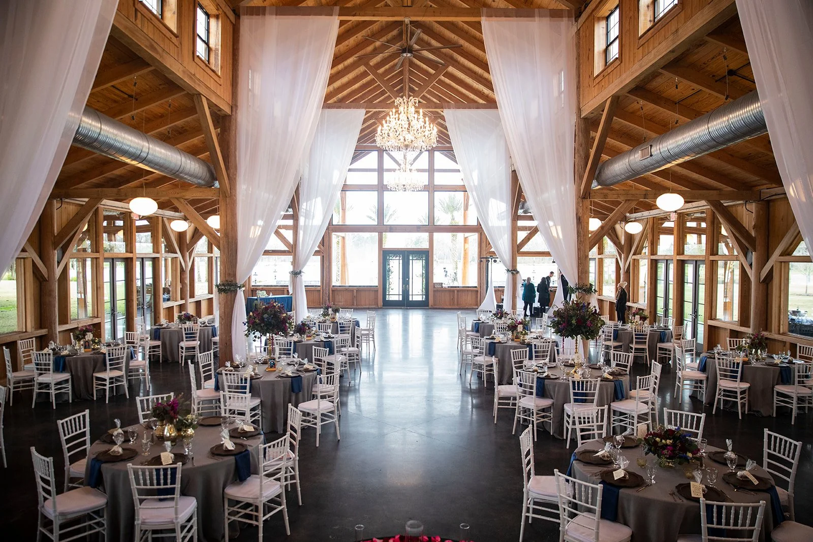 Elegant barn wedding reception venue with round tables, white chairs, floral centerpieces, draped fabric ceiling, and a chandelier, featuring large windows and wooden beams.