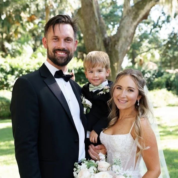 A wedding couple smiling with a young child outdoors, with the groom in a black tuxedo and the bride in a white dress, holding flowers.