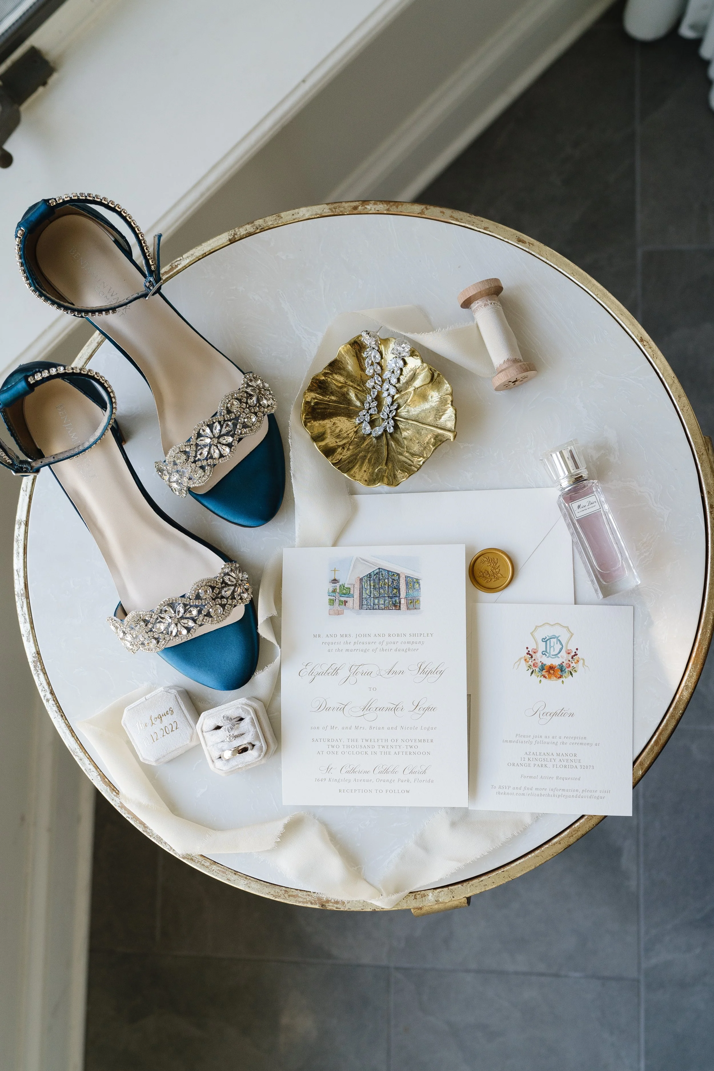 Overhead view of bridal accessories on a round white table. Items include blue jeweled sandals, a wedding invitation, a reception card, a perfume bottle, a ring box, bridal rings, a gold dish with jewelry, a ribbon spool, and a gold seal.