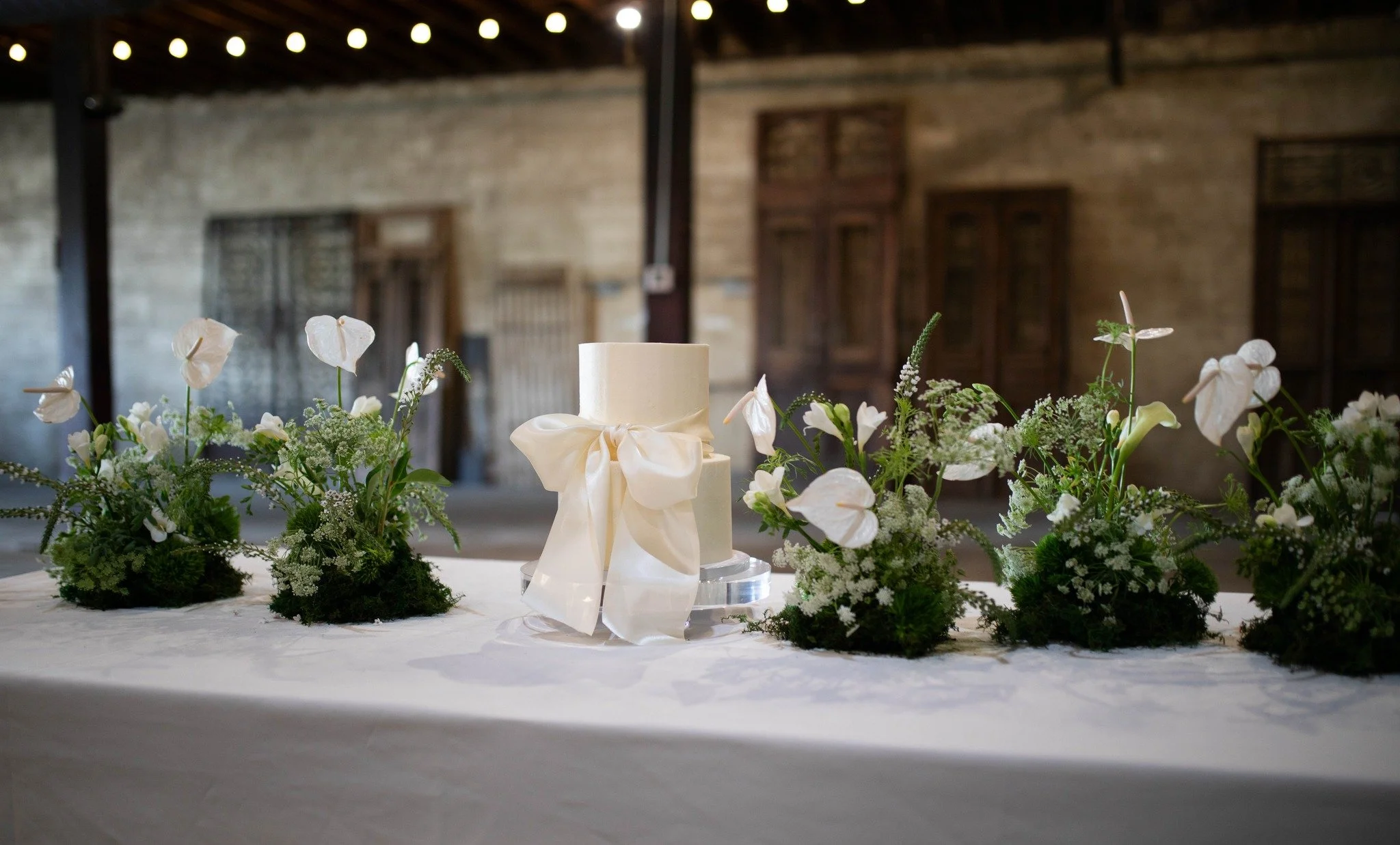 A white wedding cake with a satin ribbon bow, surrounded by white flowers and greenery, on a table with a white tablecloth. In the background, rustic wooden walls and windows are visible.