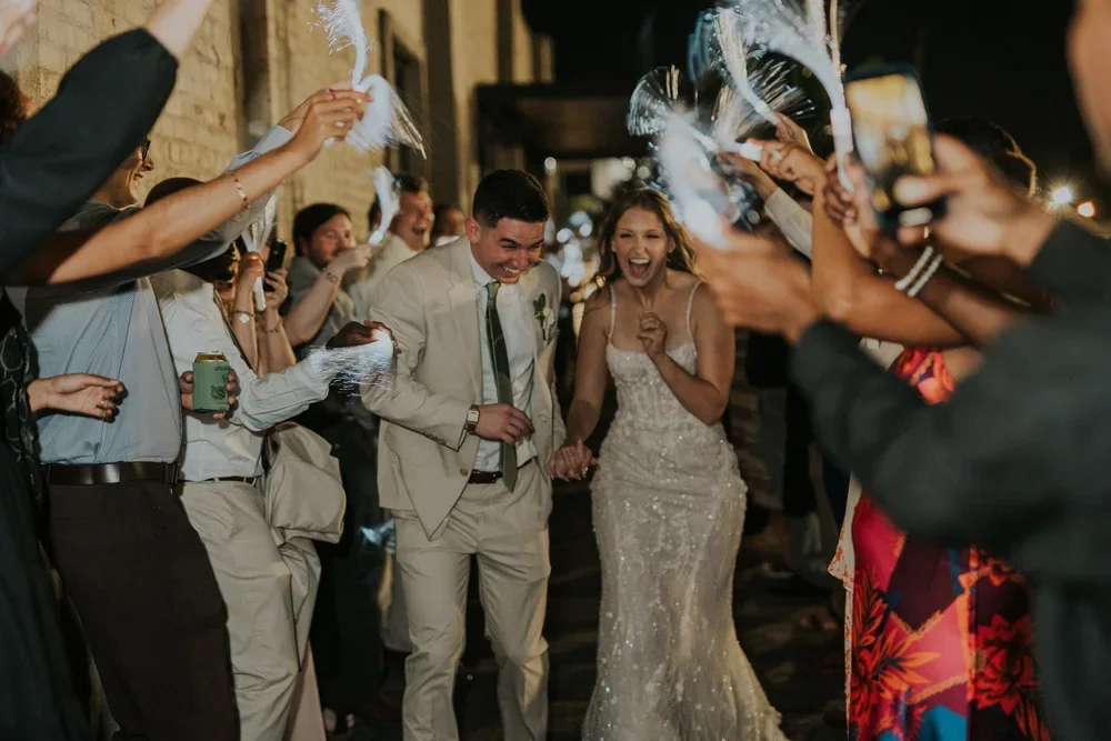 A bride and groom walking through a crowd of friends and family at their wedding reception, with guests holding up feathers and taking photos.
