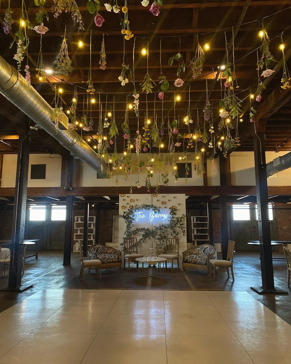 Indoor event space decorated with hanging flowers and string lights. The area features elegant seating and a backdrop with a neon sign that reads 'The Benzs', suggesting a wedding or celebration setup.